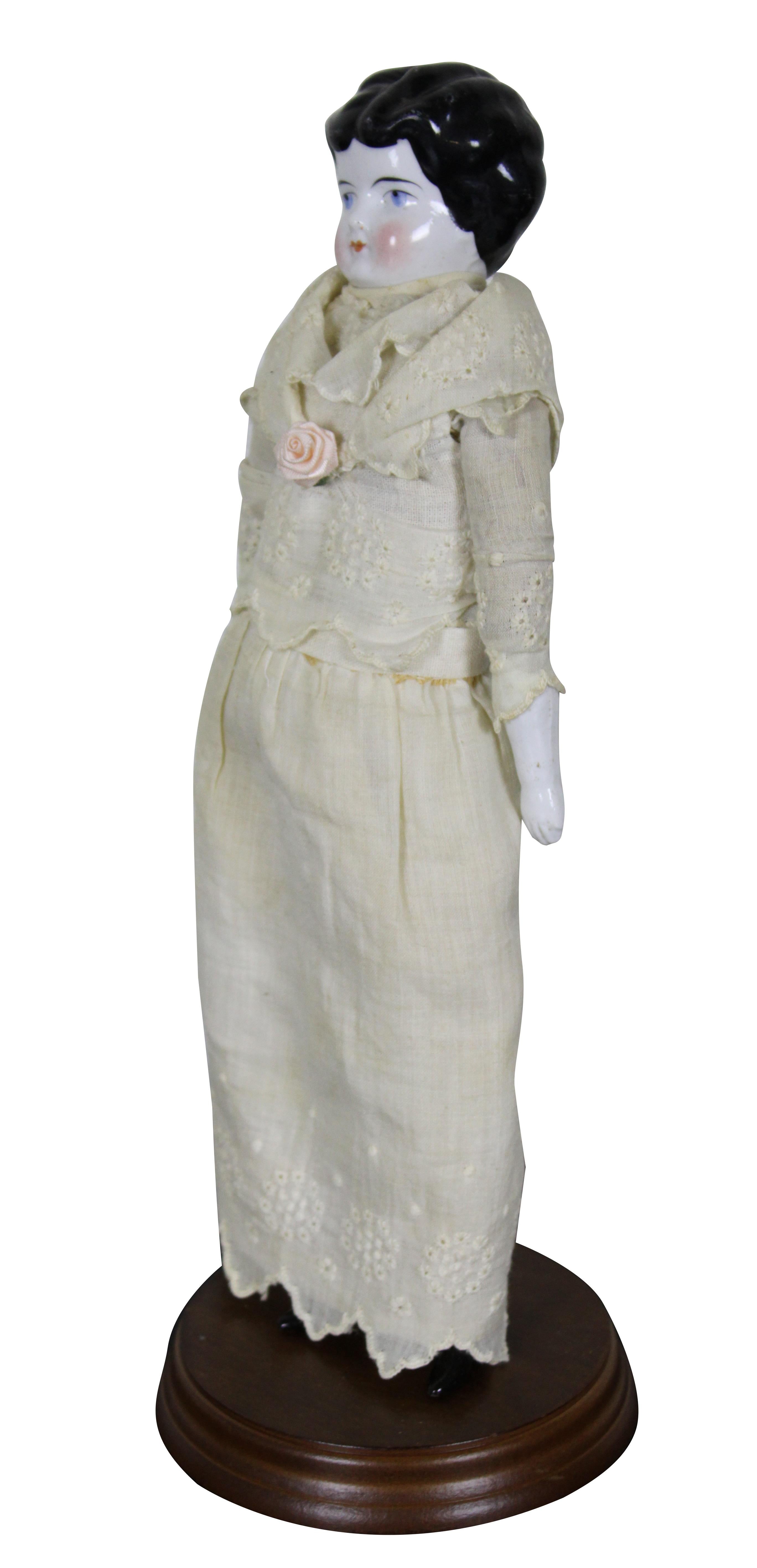 Antique 19th century German china head doll with black hair and blue eyes, dressed in a well kept lace dress with rose; displayed on a contemporary doll stand. Measure: 11”.
 