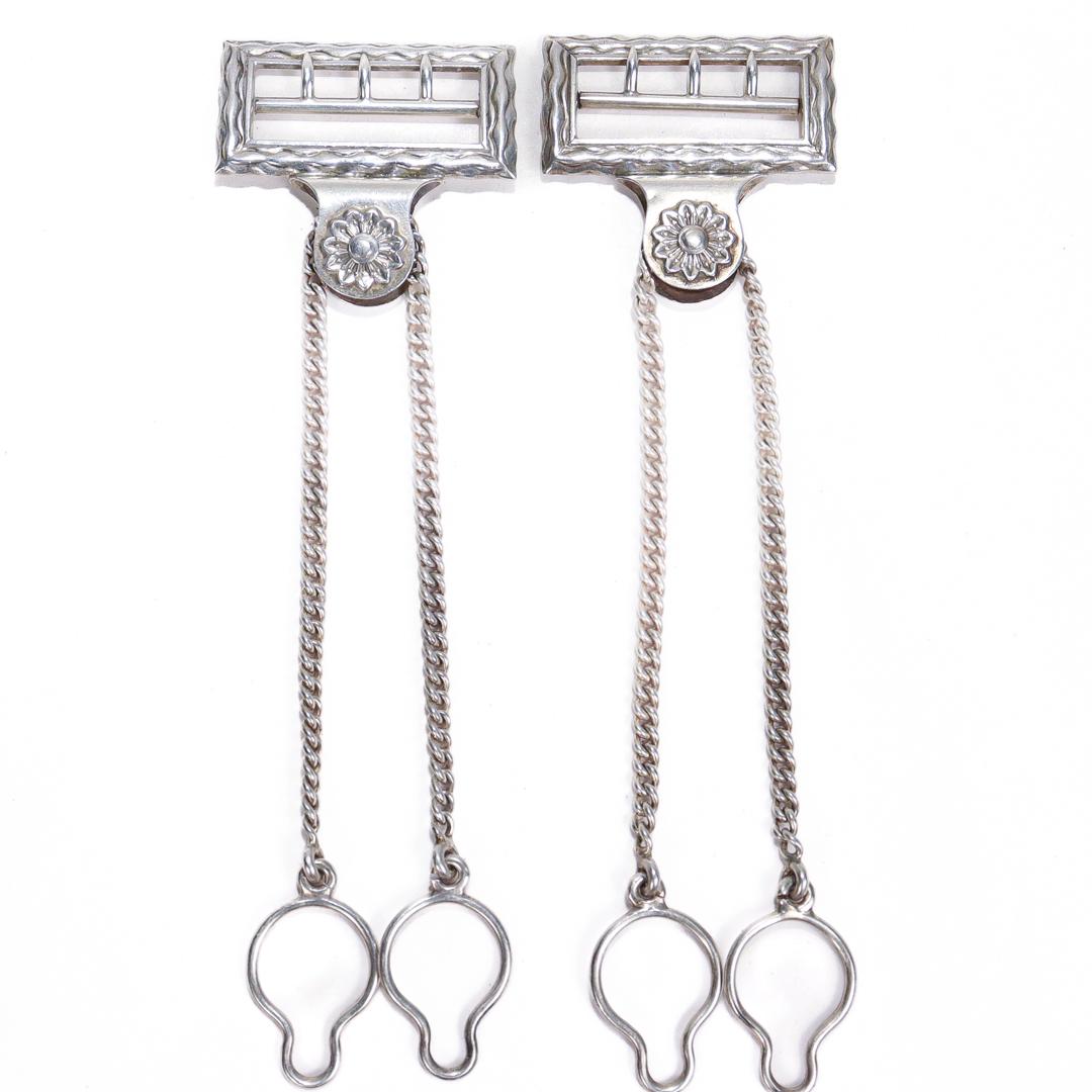A fine pair of antique silver suspender buckles

In sterling silver.

By Gorham Manufacturing Co.

Each buckle with a wavy pattern around its edges and an embossed flower where chain and buckle meet.

One buckle is monogrammed to the reverse for