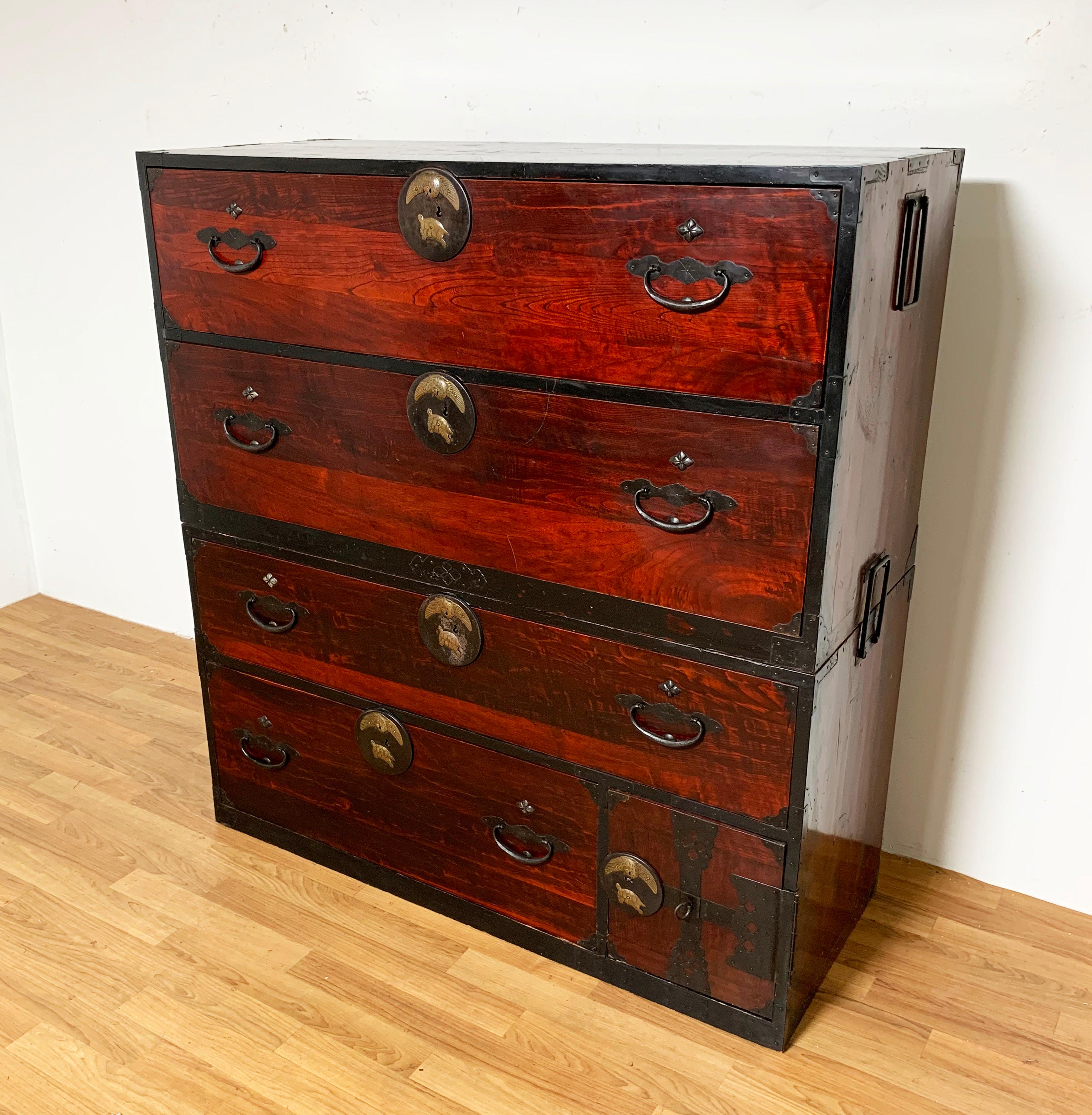 An antique 19th century Japanese Meiji period two piece tansu chest in red lacquer and akamatsu wood with hand forged iron hardware and applied brass “Tsuru-Kame” decoration depicting a crane and tortoise. These symbols are considered among the