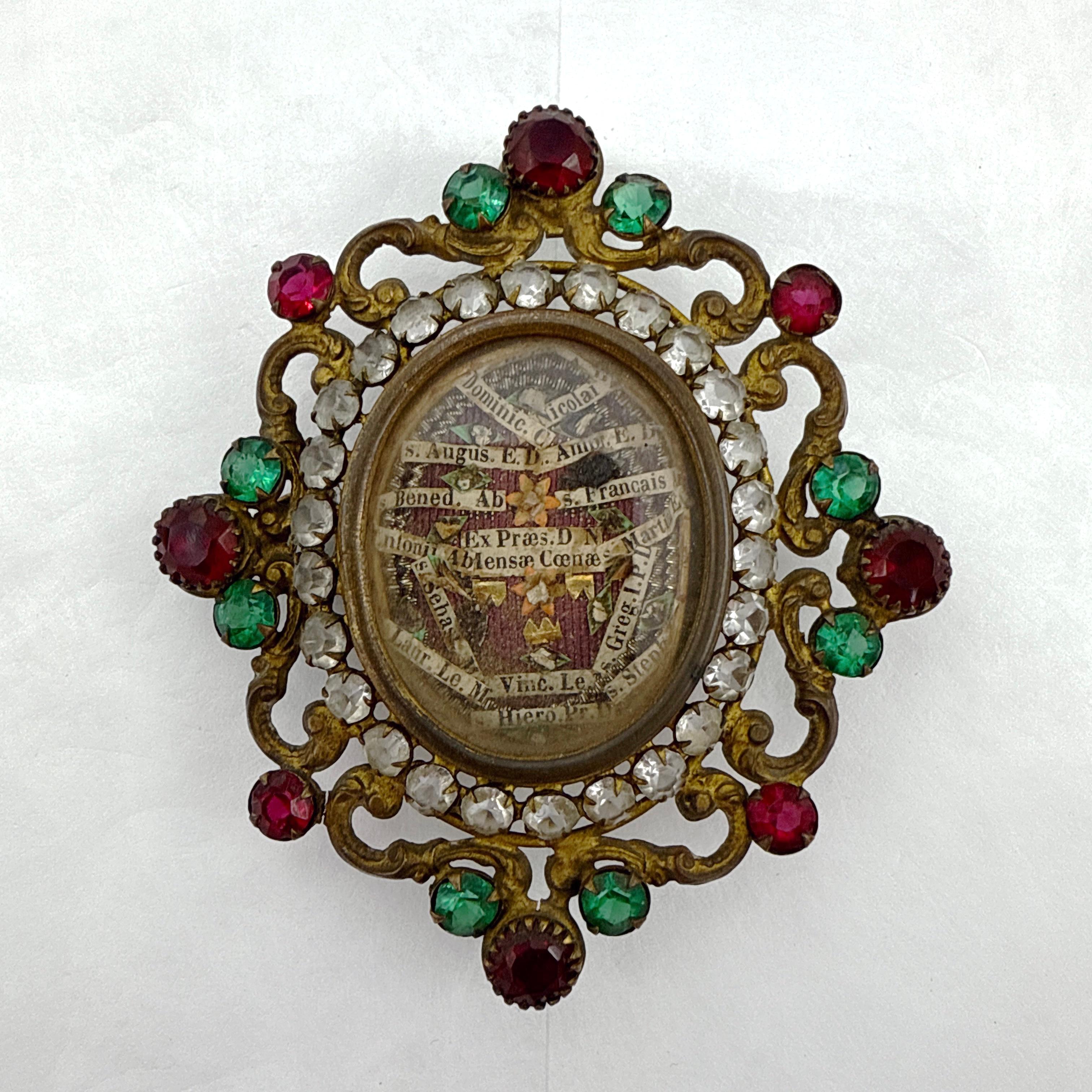 Un reliquaire catholique romain du XIXe siècle finement ouvragé présentant de multiples reliques de saints conservées sous verre dans un cadre en métal doré. Le cadre orné est décoré de pierres en pâte rouge, verte et claire dans un motif floral