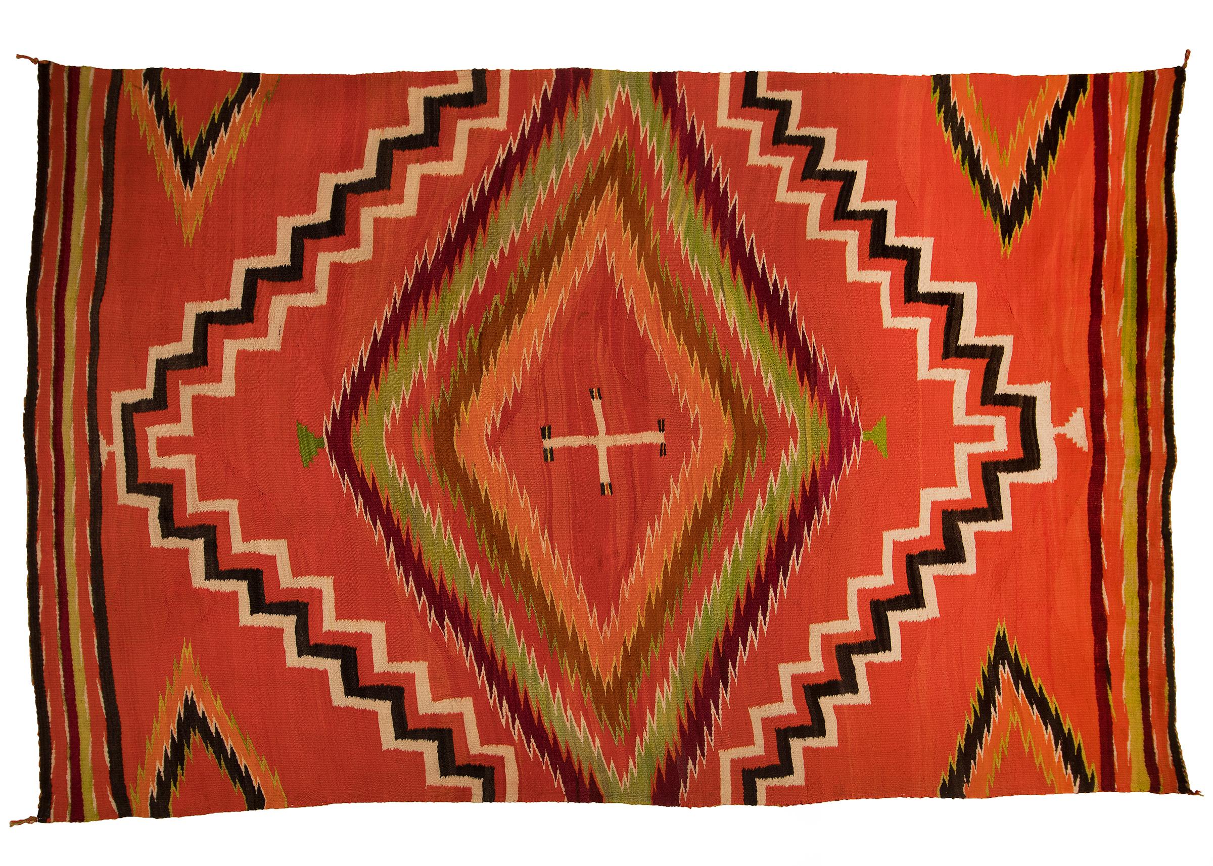 A vintage 19th century Navajo Textile in a wearing blanket format with a soft, fine weave of native hand-spun wool in aniline red, orange and green with natural fleece colors of ivory and brown/black. A central cross is surrounded by diamond