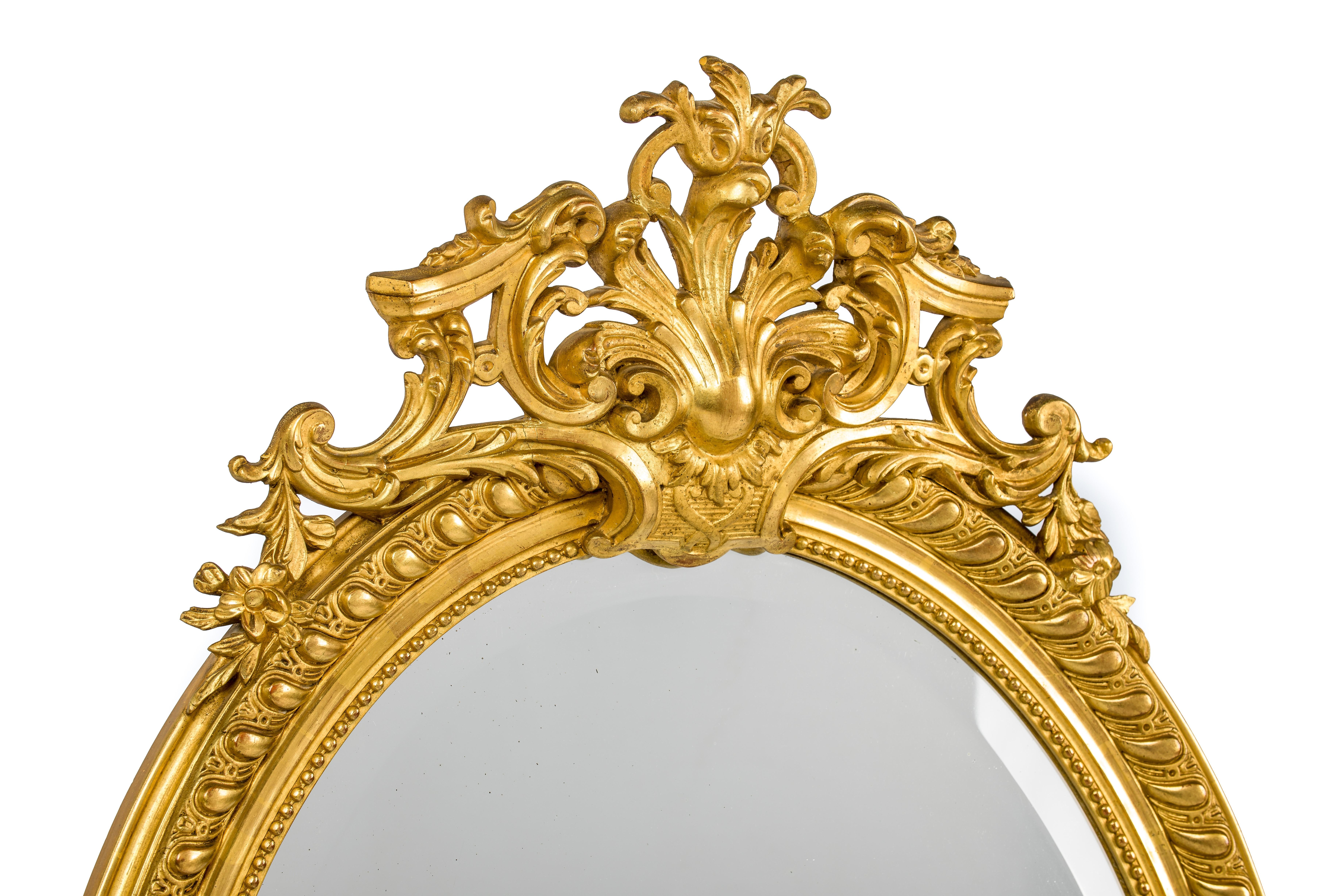 This beautiful antique gold leaf gilt oval mirror was made in southern France in the late 19th century. The top ornament is centered upon an acanthus pediment flanked by C-scrolls, acanthus, and floral elements. The lower ornament has the same
