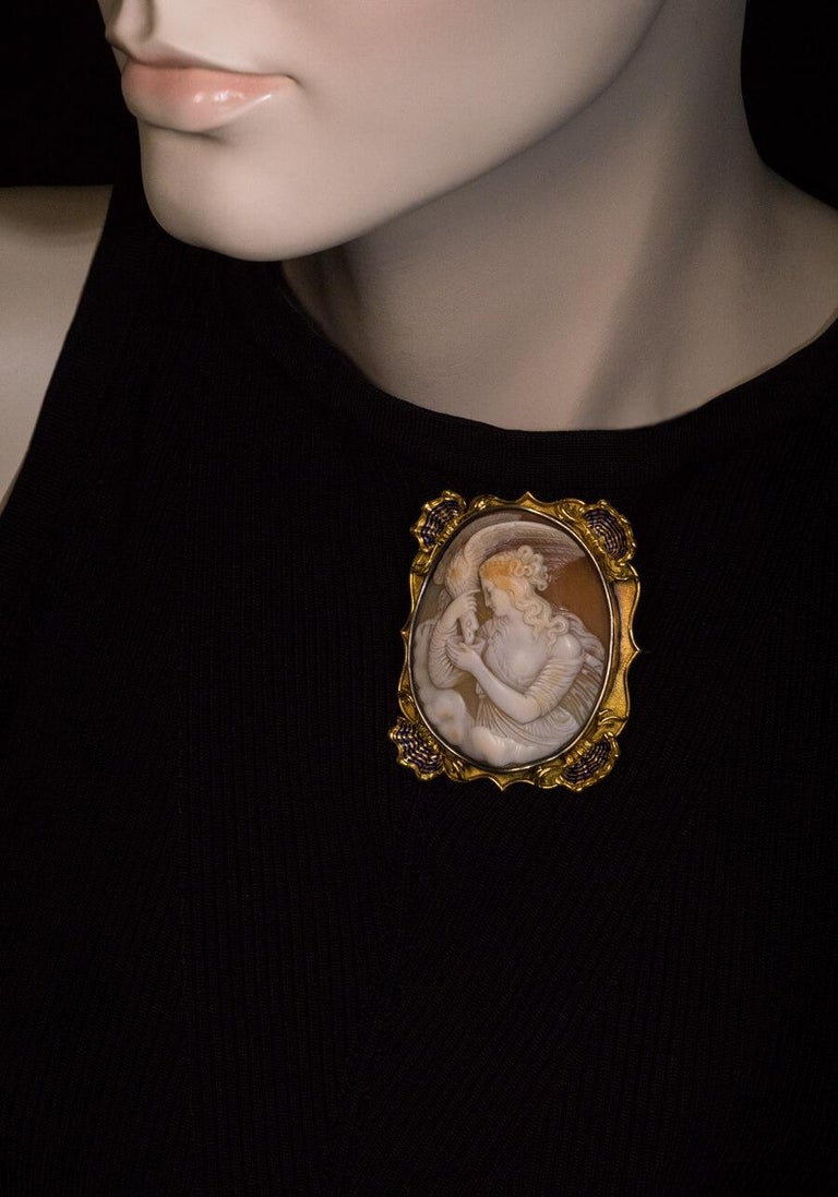 Italian or French, circa 1860
A large finely carved shell cameo of Hebe and the eagle of Zeus is set in an ornate 18K gold bezel and mounted as a brooch. The corners of the bezel are accented by gold shells embellished with blue enamel.
Hebe, the