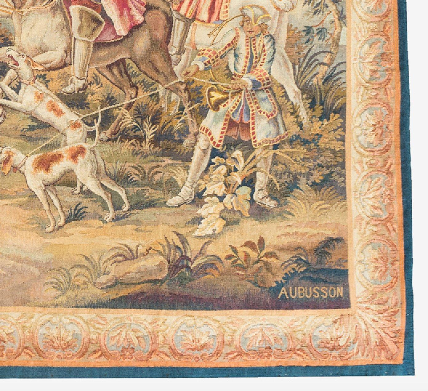 Ancienne tapisserie d'Aubusson du XIXe siècle représentant une scène de chasse sur les rives d'une rivière avec des hommes sur des chevaux blancs et des chiens de chasse qui ont capturé un cerf dans la rivière. La scène est entourée de terrains