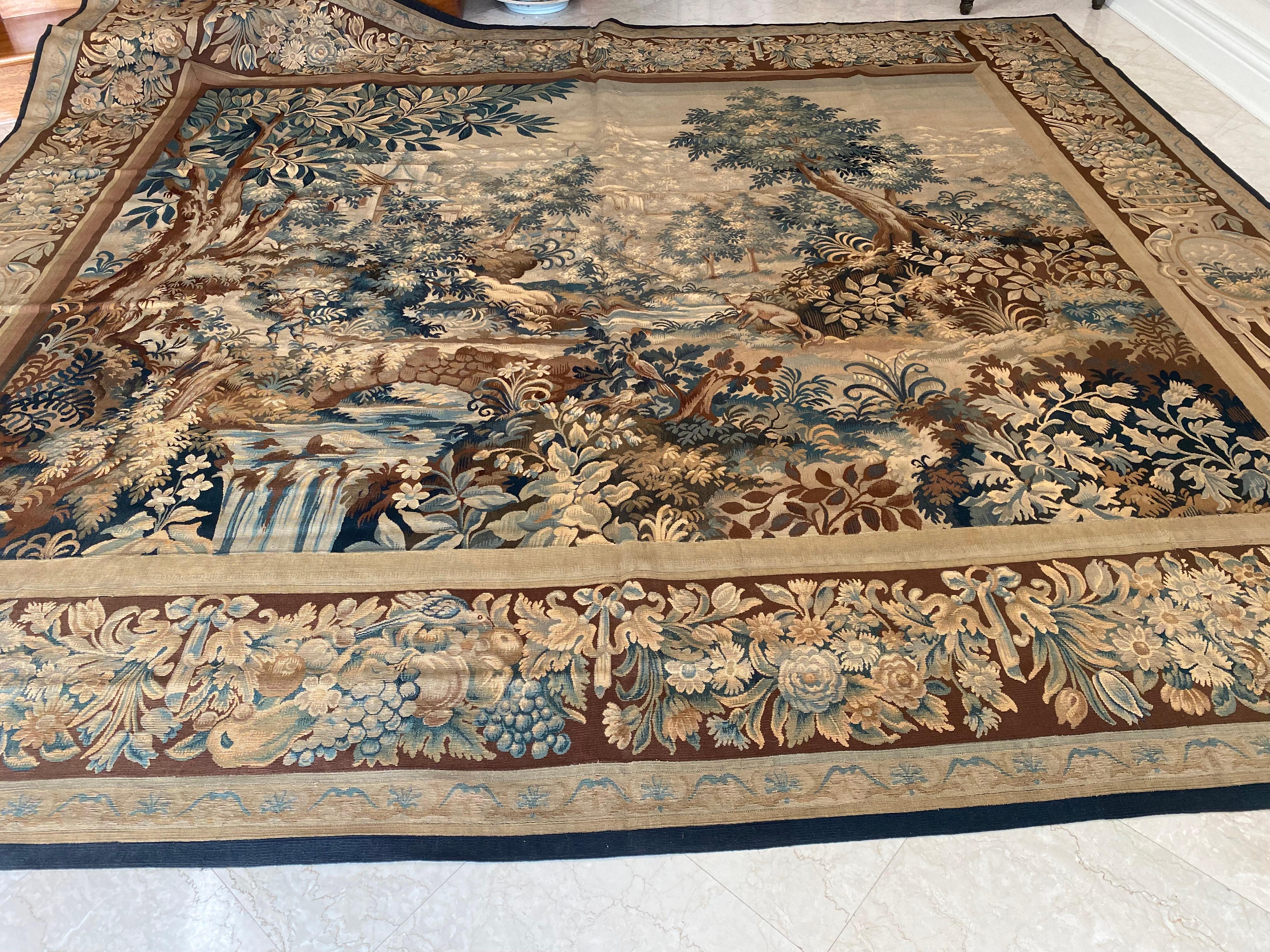This is a lovely antique 19th century square green French Verdure tapestry depicting a beautiful summer scene of a countryside with lush trees and vegetation, a river with a stone bridge, and a path leading to a manor in the distance. It has an