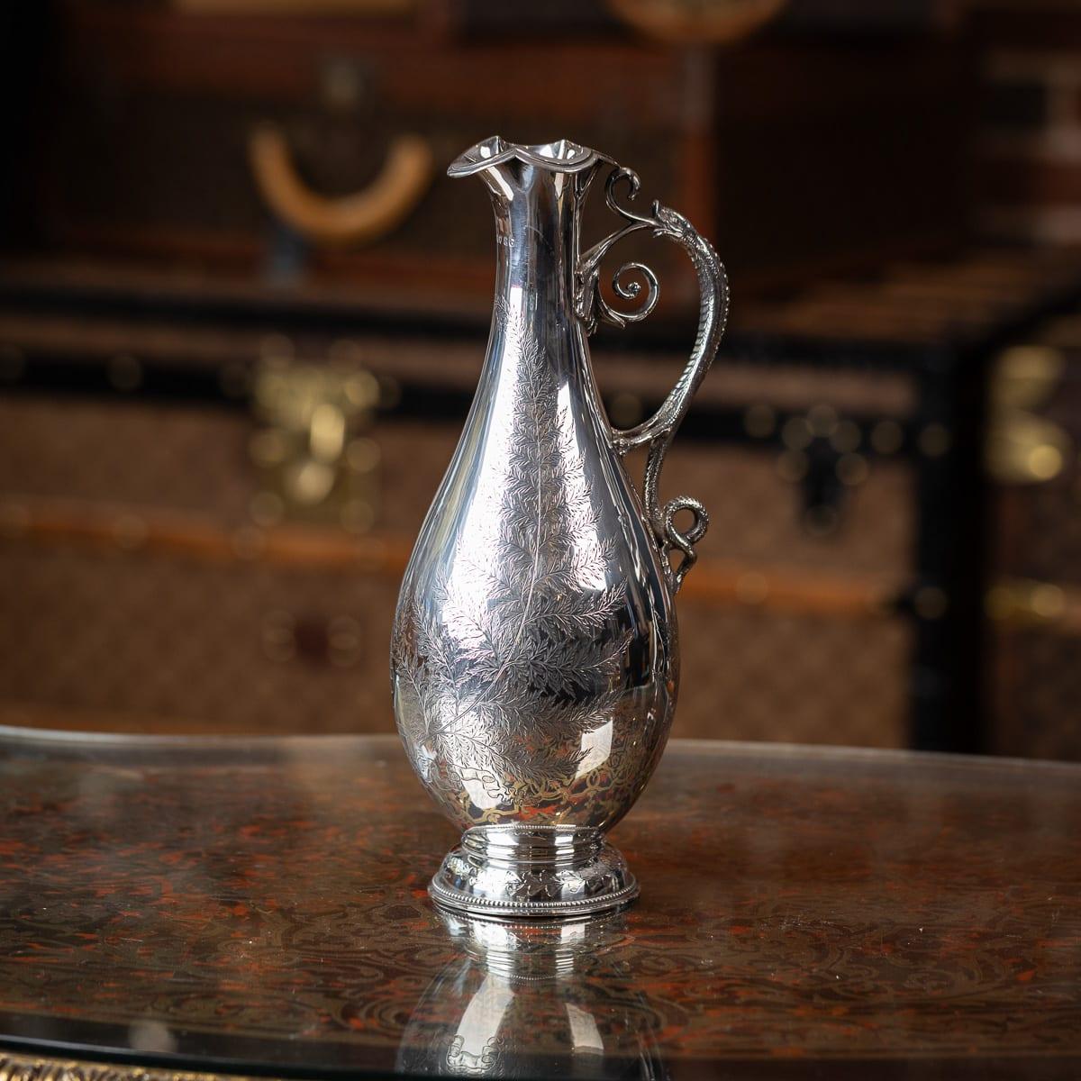 Ancienne aiguière victorienne en argent massif du 19e siècle, présentant un corps bulbeux et effilé avec un décor de feuillage et une poignée en forme de lézard. Reposant sur une base ronde et évasée avec des motifs de feuillage assortis, cette