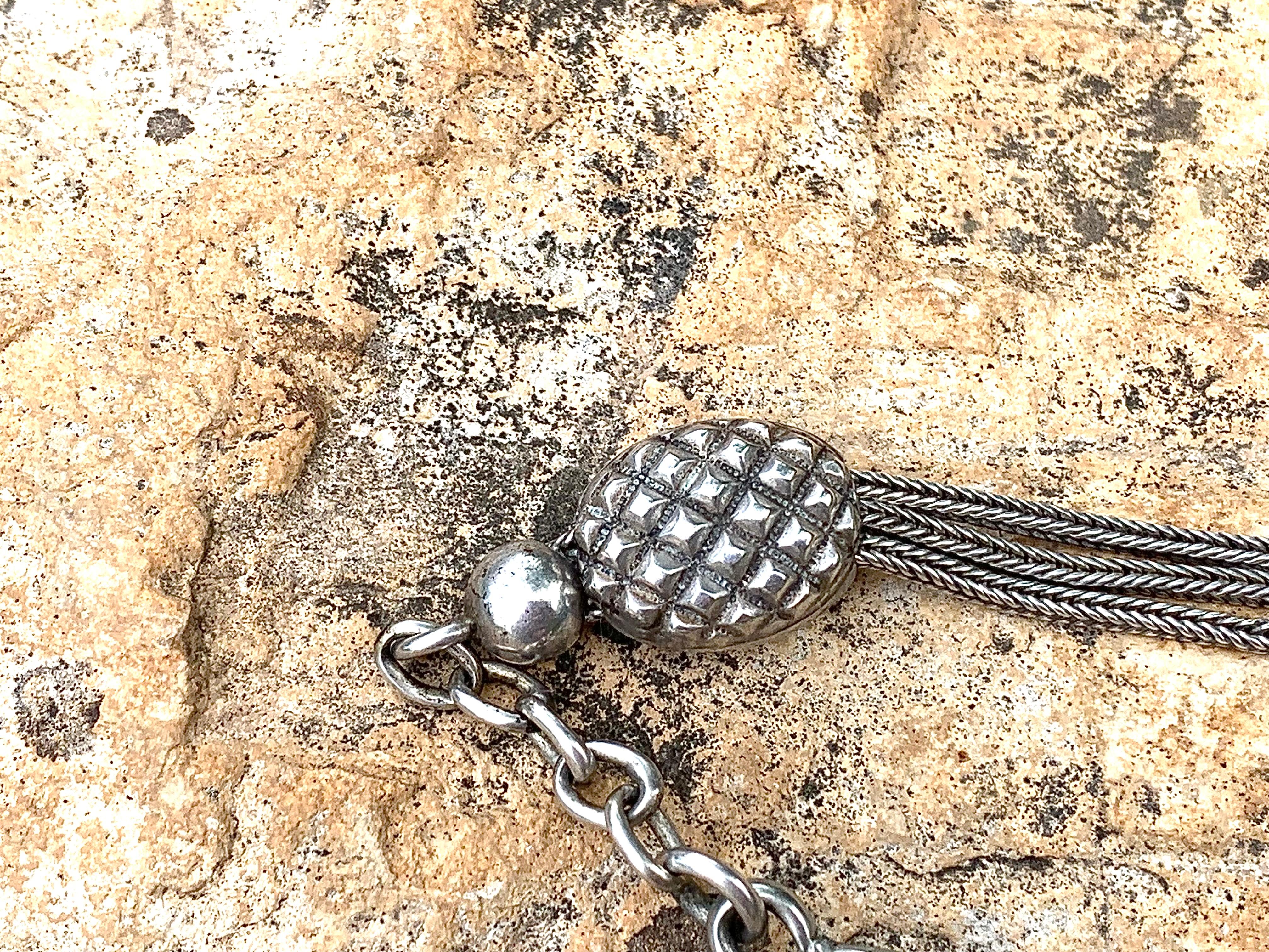 Cette chaîne de montre en argent a été fabriquée dans le dernier quart du 19e siècle. Il est composé de chaînes en argent à queue de renard lisse, avec deux glissières à la surface d'une pomme de pin stylisée. Un œillet porte un poinçon