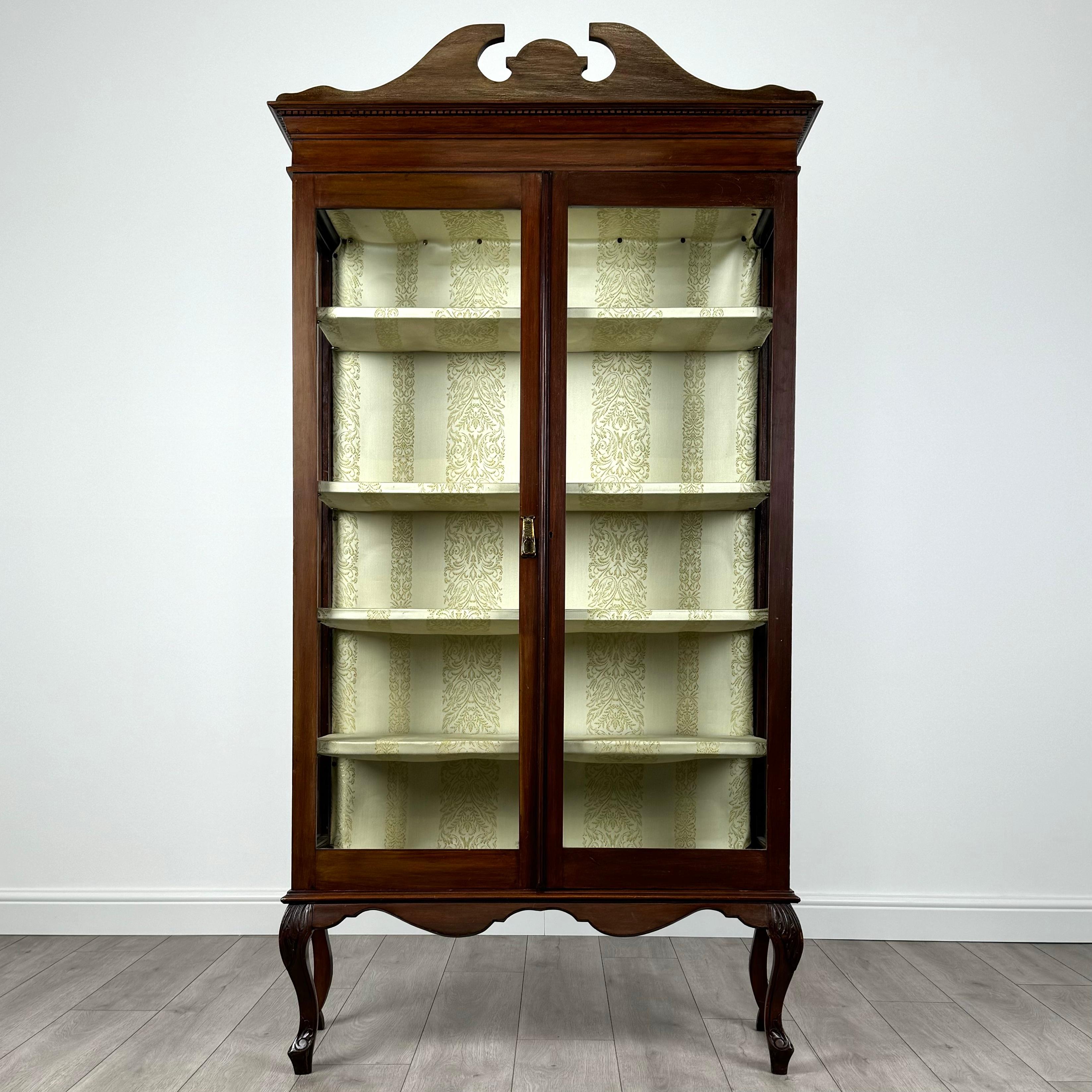 Cette élégante vitrine antique du XXe siècle, fabriquée en acajou, est une pièce raffinée qui allie fonctionnalité et design classique. Le meuble est gracieusement élevé sur des pieds cabriole, chacun orné de genoux magnifiquement sculptés, qui