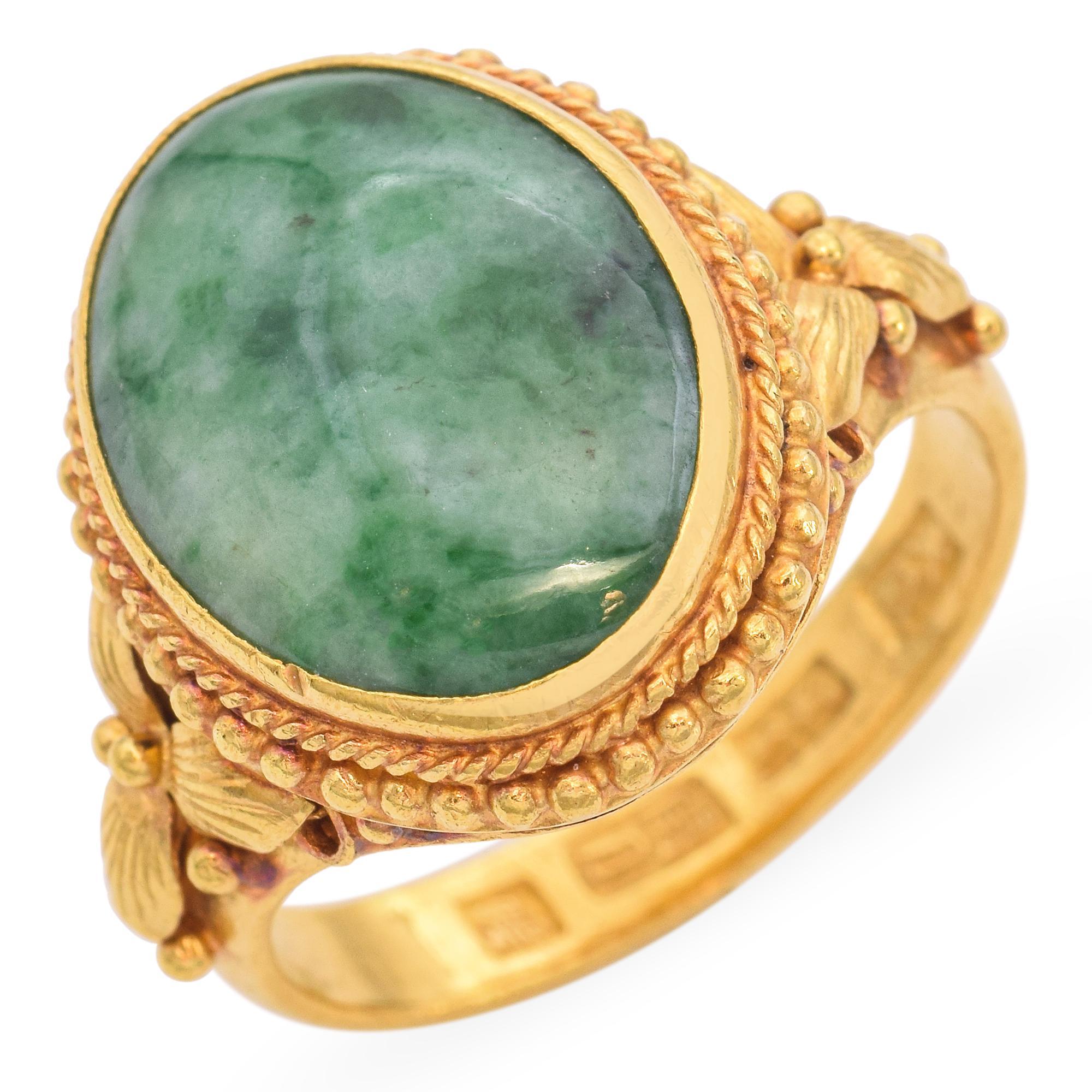 An elegant ring featuring a large oval jade, set in a detailed gold band. The stone displays a rich, deep green color with natural variations, adding depth and character. It is encased in an intricate gold setting with rope-like detailing around the