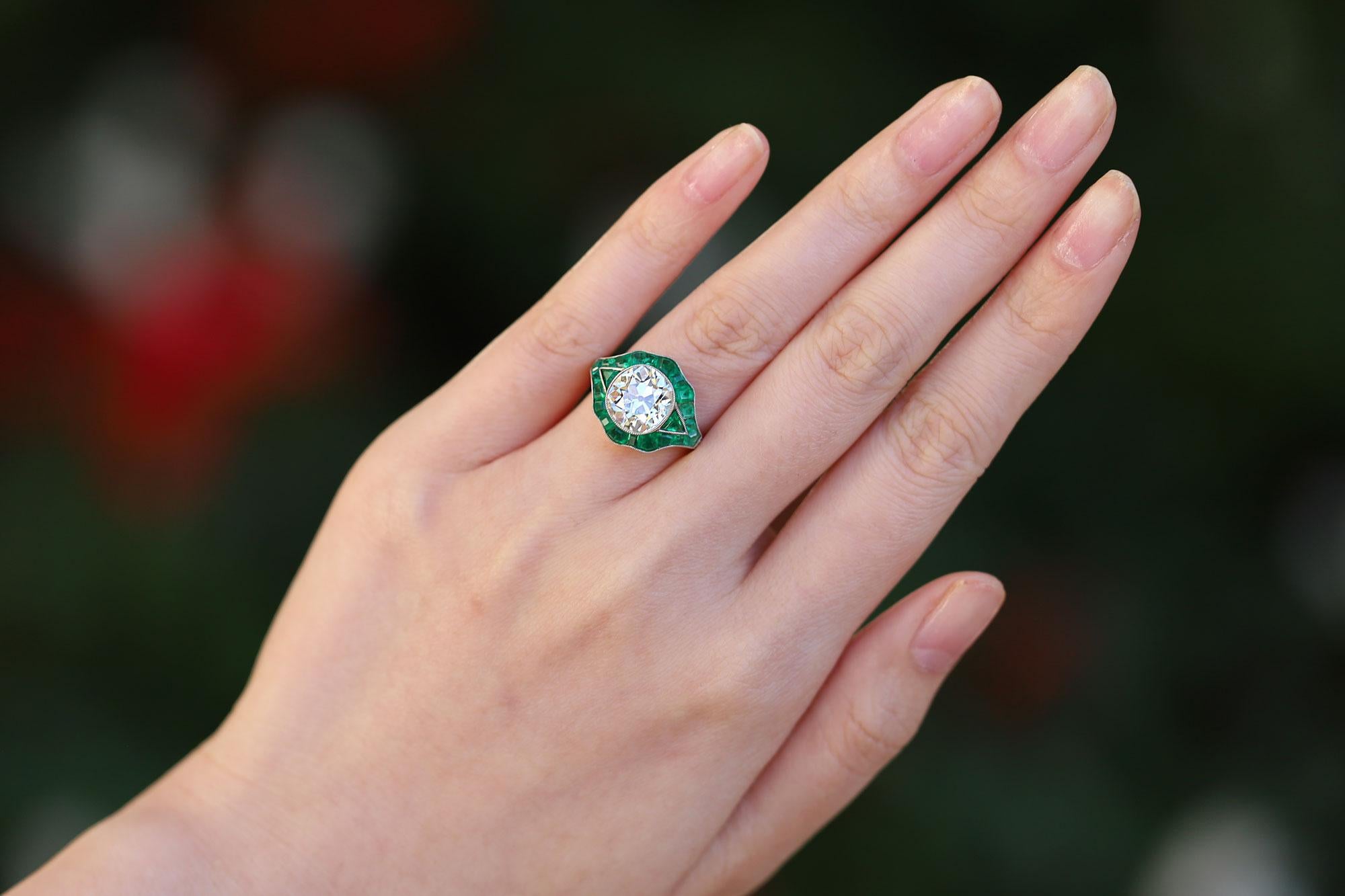 Centering on a large 3.05 Carat, chunky and scintillating antique old mine cut diamond, this Art Deco inspired engagement ring is a show stopper. Encased in a platinum, milgrain-studded bezel and surrounded with rich, green emeralds, this important