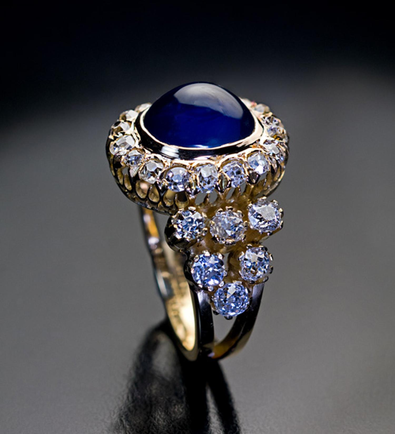 Circa 1890

A Victorian era antique 18K gold ring is centered with an oval 5.37 ct cabochon cut natural sapphire of a velvet dark blue color. The sapphire is framed by 18 sparkling old mine cut diamonds. The split shoulders of the ring are prong-set