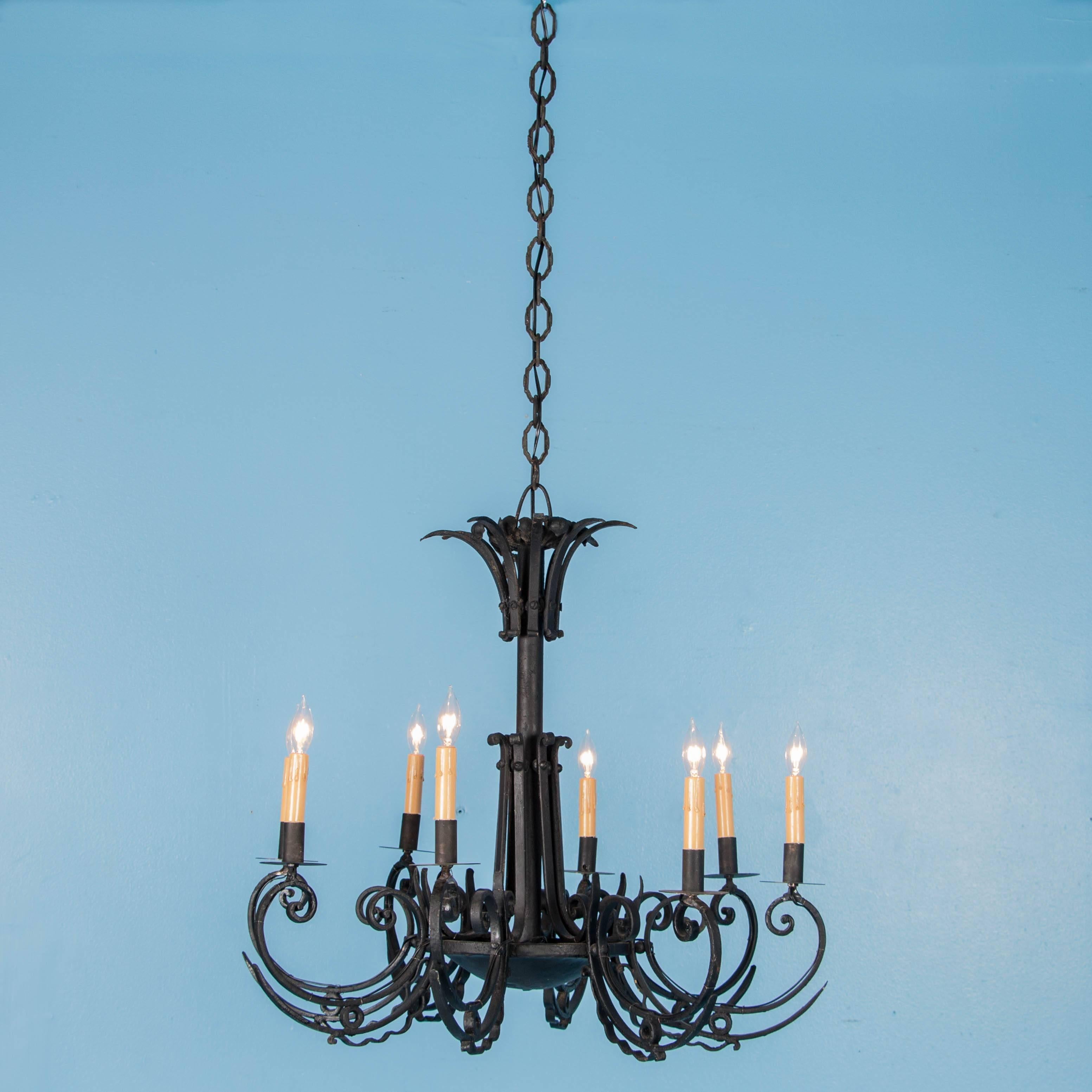 This antique French chandelier (circa 1920), features eight ornate wrought iron arms, each set with a single light and a candle cover resting on a drip pan. Notice how below each of the scrolled arms there are 2 layers of curved iron, one spiked and