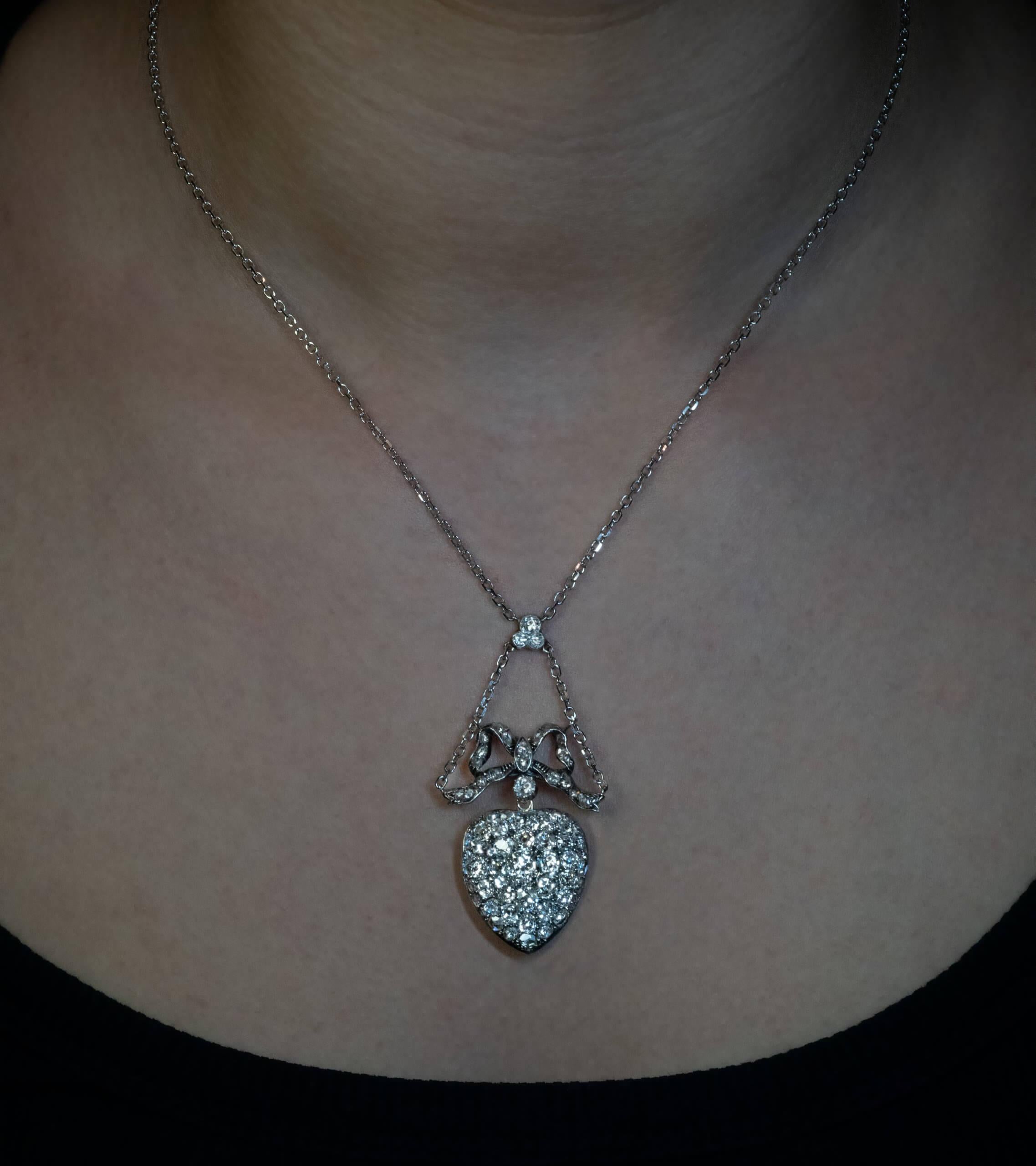 Circa 1870  The necklace is designed as a heart locket pendant surmounted by a ribbon bow. Both are finely crafted in silver topped 14K gold (front – silver / back – gold). The 14K white gold chain and detachable bail are modern.  The heart pendant