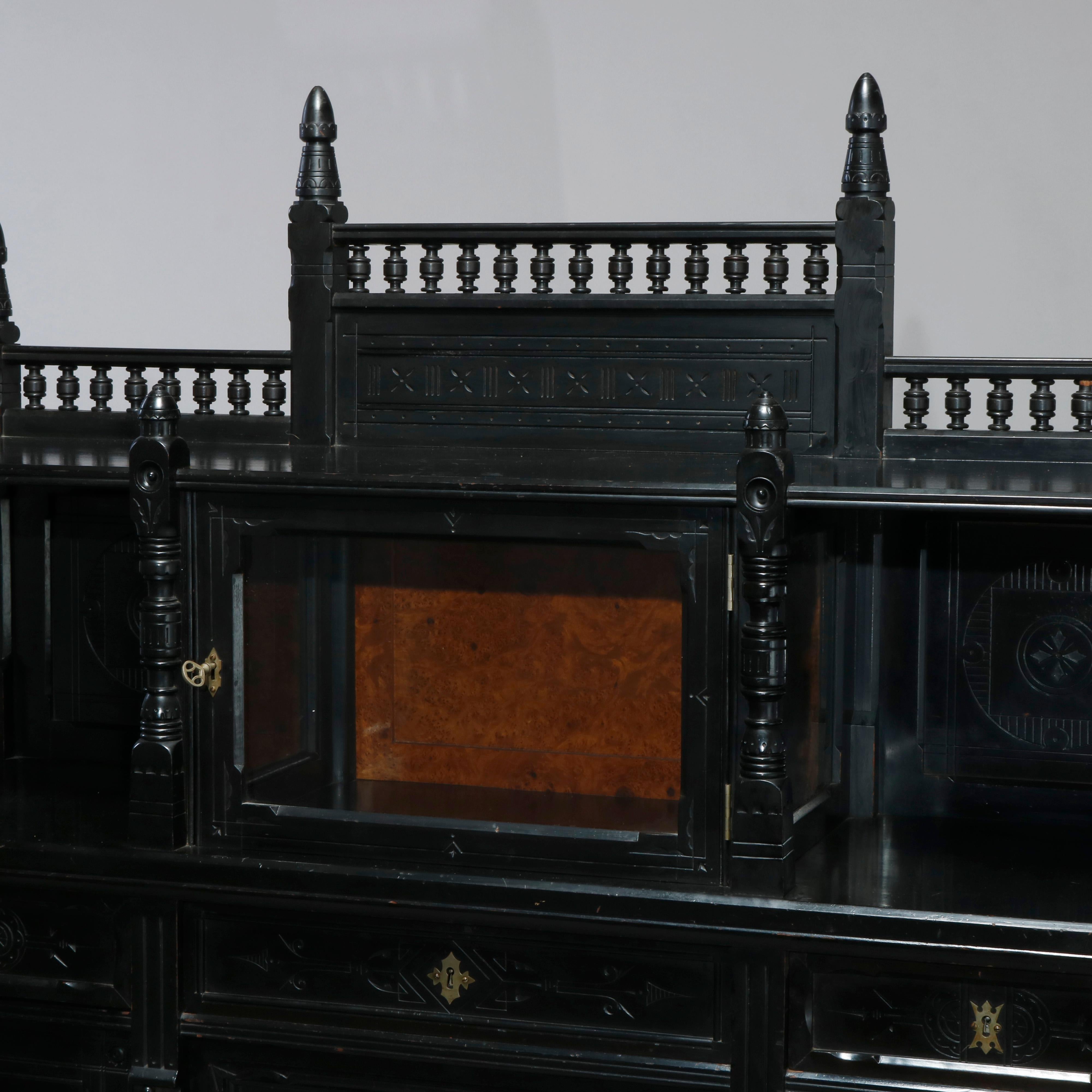An antique Aesthetic Movement credenza in the manner of Kimbel & Cabus offers ebonized cabinet with spindled rail surmounting floral and foliate incised upper having central locking vitrine with glass door opening to burl back display with flanking