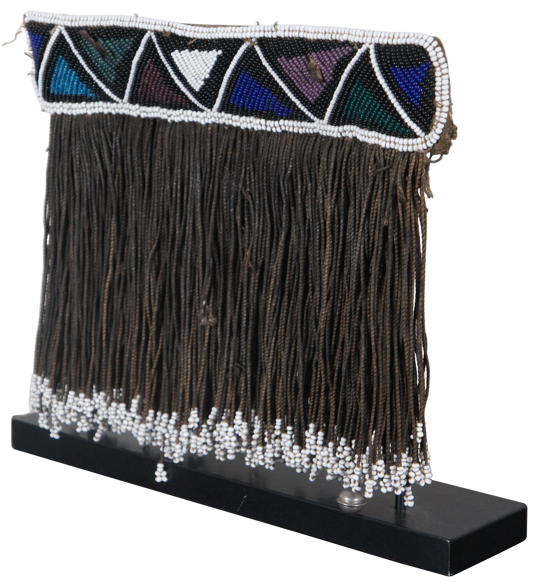 Antique beaded art featuring a multi-color chevron patterned panel with long beaded fringe and one bell displayed on Iron stand.

Measures: 12.5” x 0.5” x 10.75” / with stand - 14” x 3.25” x 12” (width x depth x height).