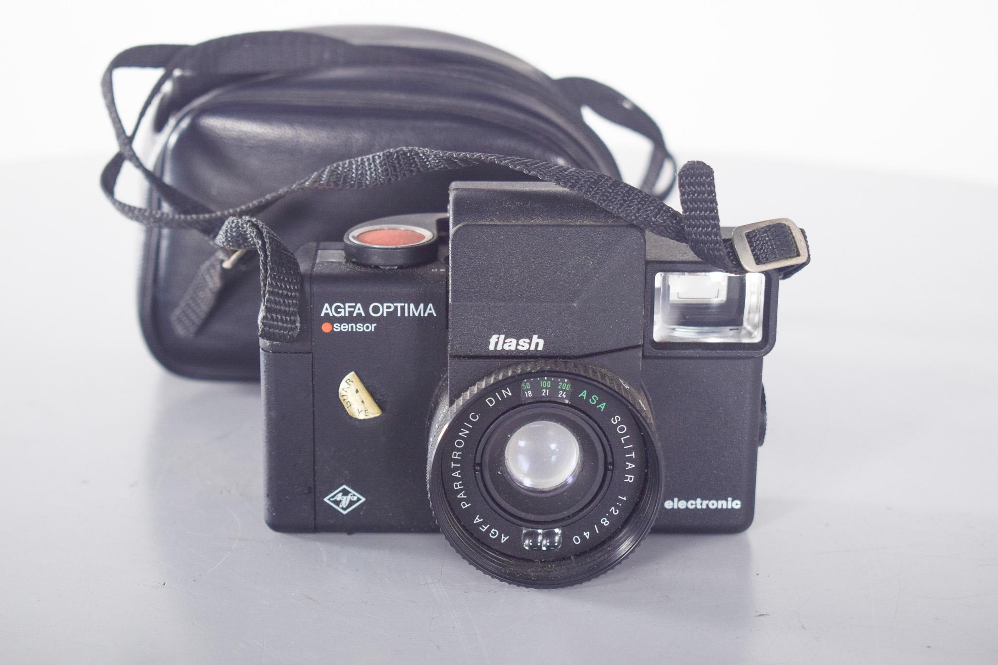 Appareil photo AGFA, Optima Flash, avec étui. Appareil photo analogique 35 mm, appartenant à la série emblématique Optima, réputée pour sa facilité d'utilisation et sa robustesse. Ce modèle présente un design compact et fonctionnel, avec un corps en