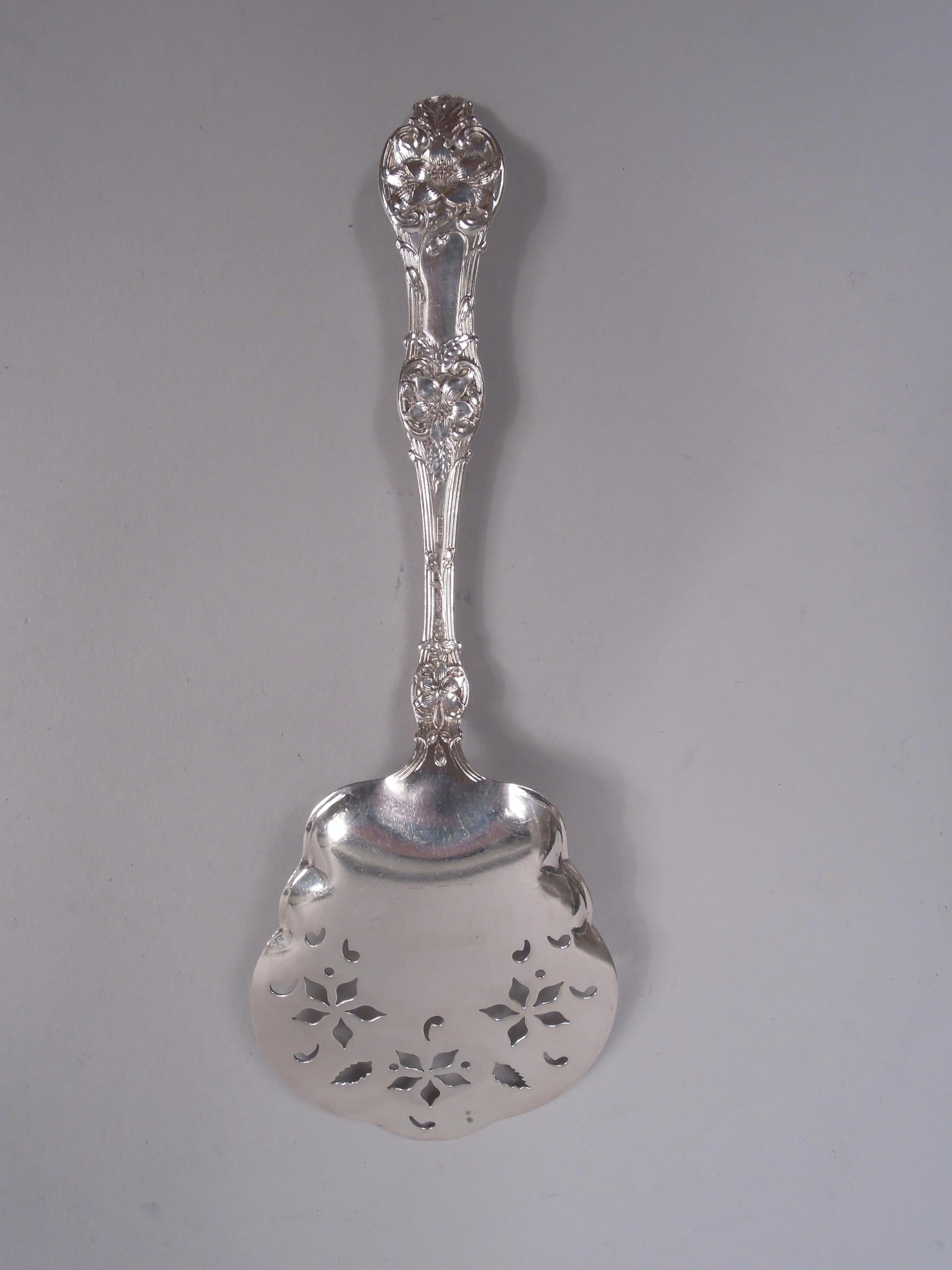 Servidor de tomates Orange Blossom de plata de ley. Fabricada por Alvin Corporation en Providence, hacia 1910. Caña cóncava y serpentina de 3 púas; ornamento floral sobre vástago enrollado y acanalado con terminal trébol. Hoja perforada y festoneada
