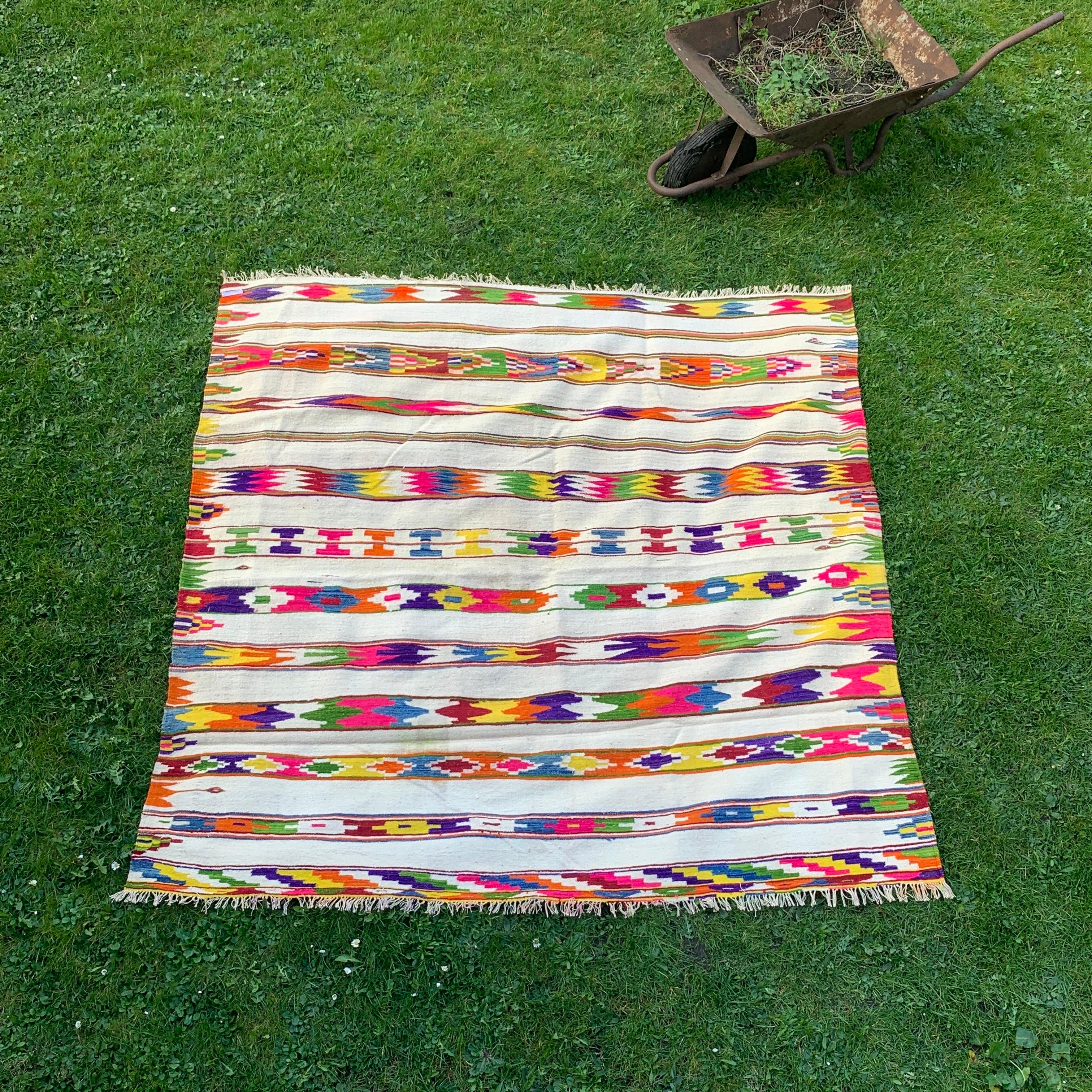 A beautiful multicoloured square rug in warm colour pallet that could be used on either can be side, making it versatile and functional. 

The base of the rug is a light beige colour, providing a neutral background for the vibrant geometric patterns