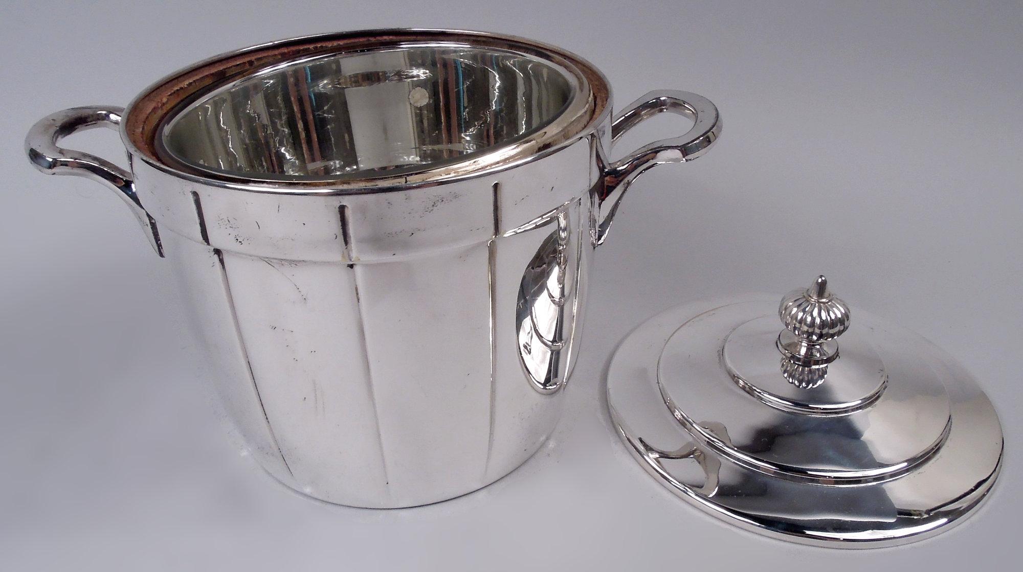 Seau à glace Art déco en métal argenté, vers 1920. Forme tonneau avec douelles ; épaule en surplomb et poignées latérales en forme d'écusson. Couvercle surmonté d'un épi de faîtage en forme de boule lobée. Doublure en verre à isolation thermique.