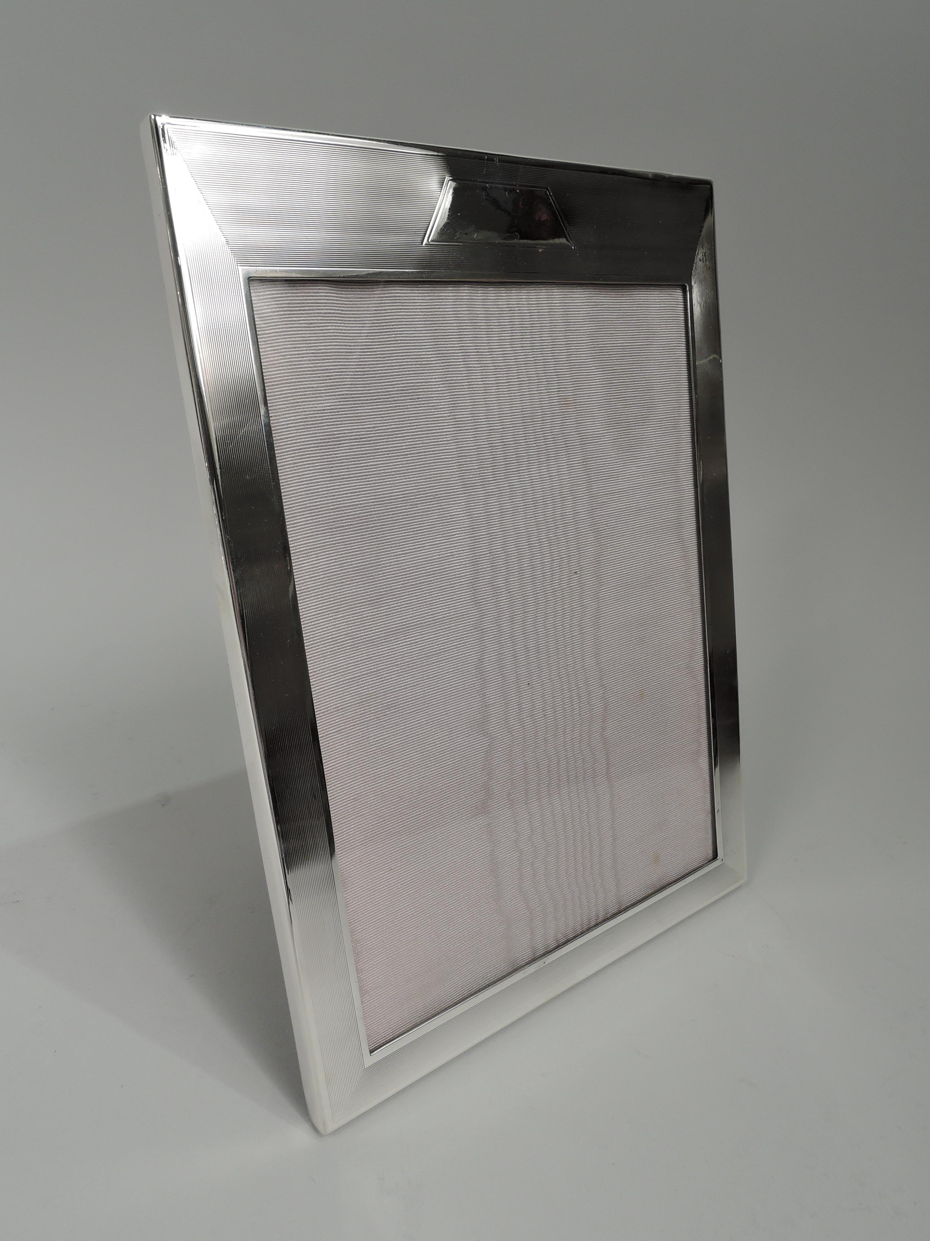 American Art Deco sterling silver picture frame, ca 1925. Rectangular window in flat surround with wraparound engine-turned lines between plain borders. Sides plain. Trapezoidal frame (vacant). With glass, silk lining, and velvet back and hinged