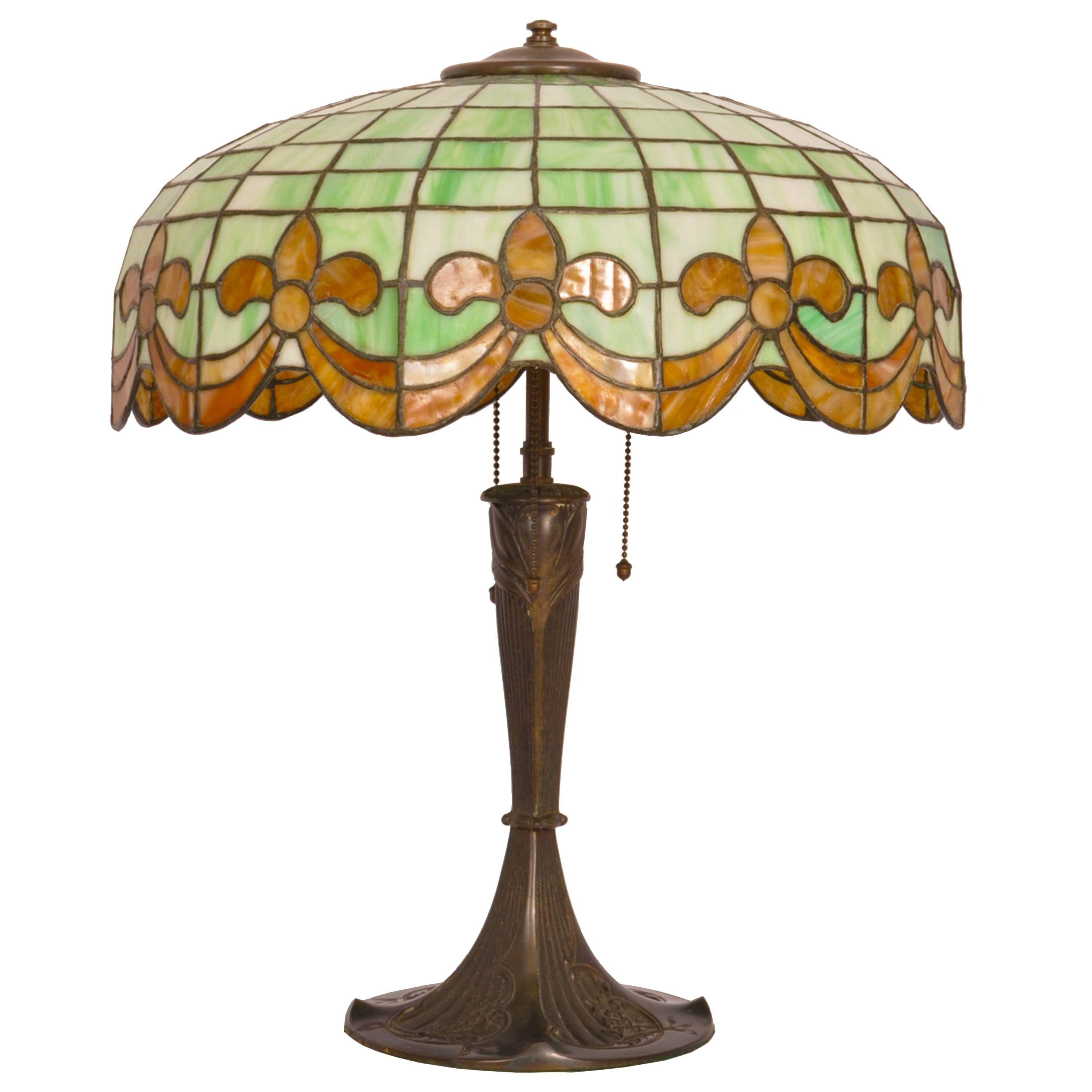 Antique American bronze and leaded glass table lamp by Wilkinson, circa 1910.
The three light lamp having a dome shaped shade with striated leaded glass green & ivory tiles and having amber colored Fleur de Lys & swagged decoration to the base of
