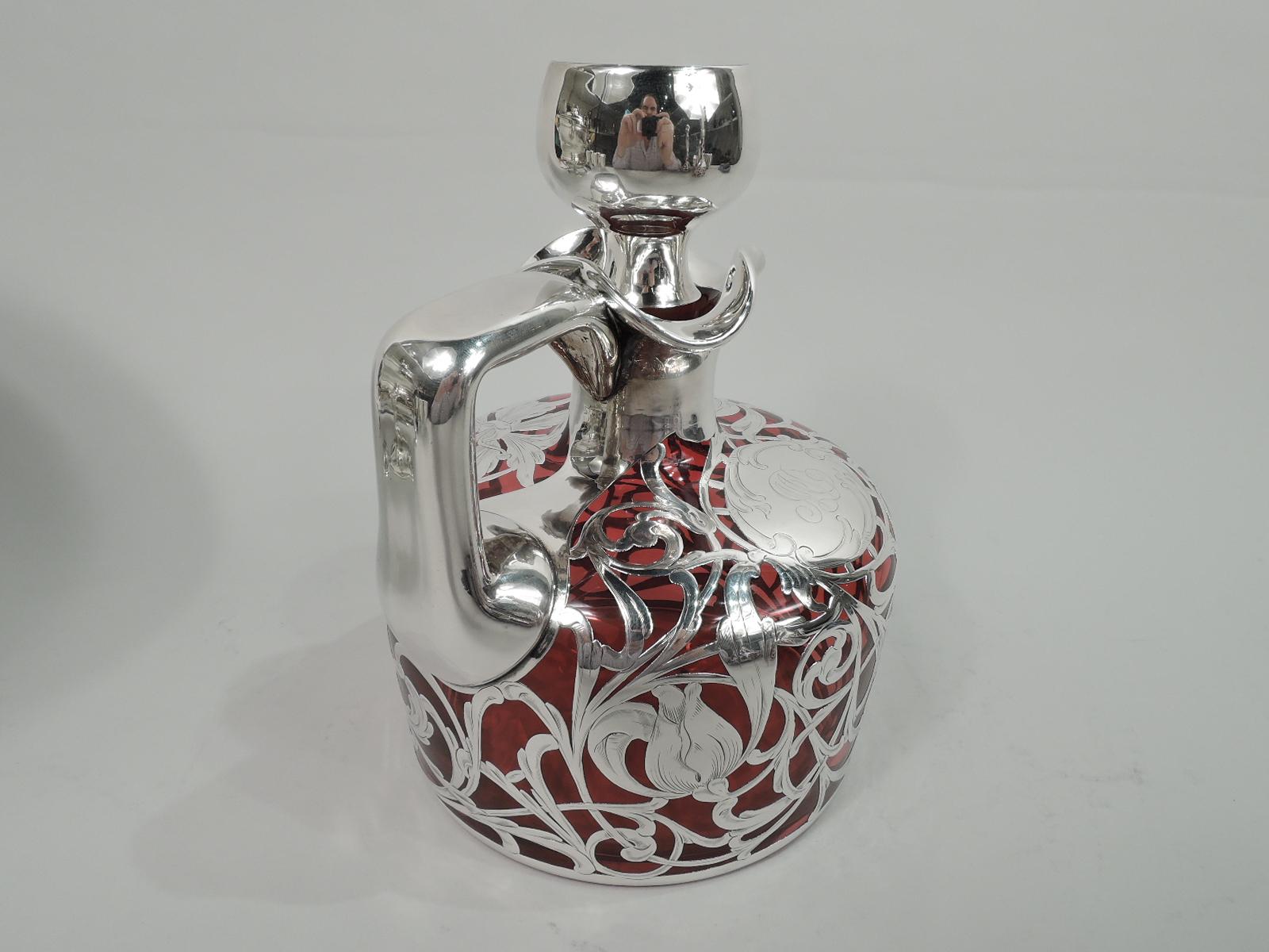 Turn-of-the-century American Art Nouveau glass jug decanter with engraved silver overlay. Drum form with curved shoulder. Neck and trefoil mouth in silver collar as is bracket handle. Flat-topped stopper same. Entwined and scrolling tendrils with