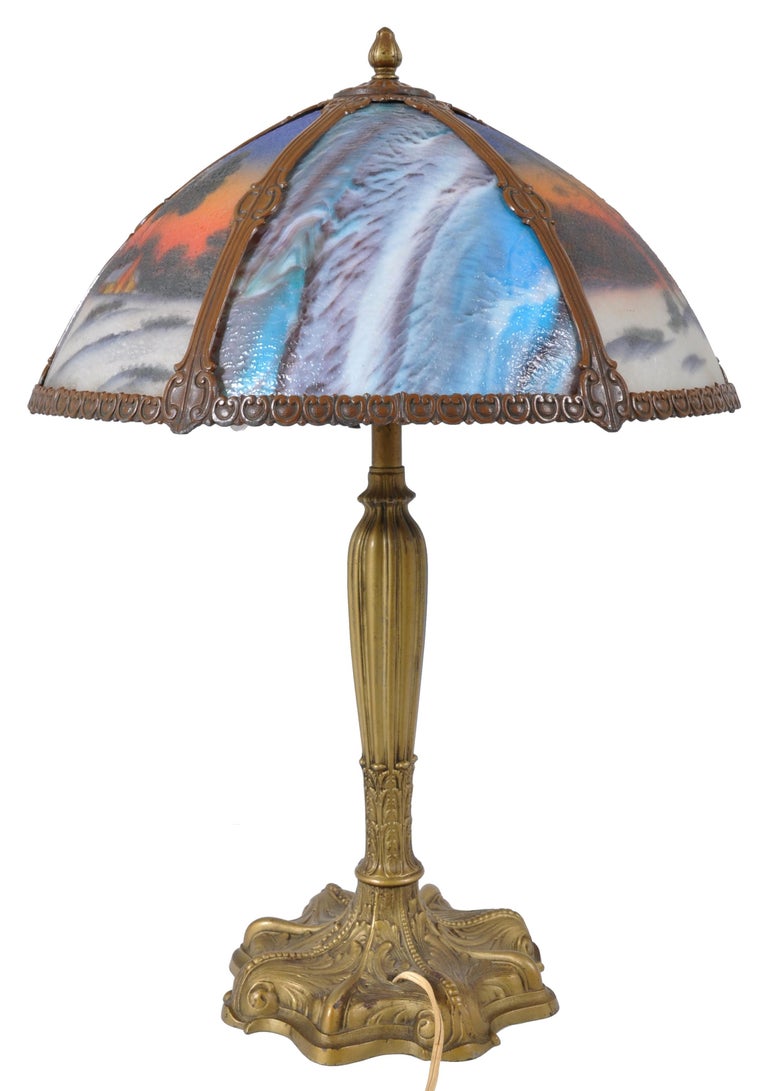 Antique American Art Nouveau reverse painted landscape table lamp, circa 1910. The Pittsburgh style lamp, possibly by Pairpoint, having a bronze base with a six-panel reverse painted landscape shade. The shade having alternating blue variegated slag