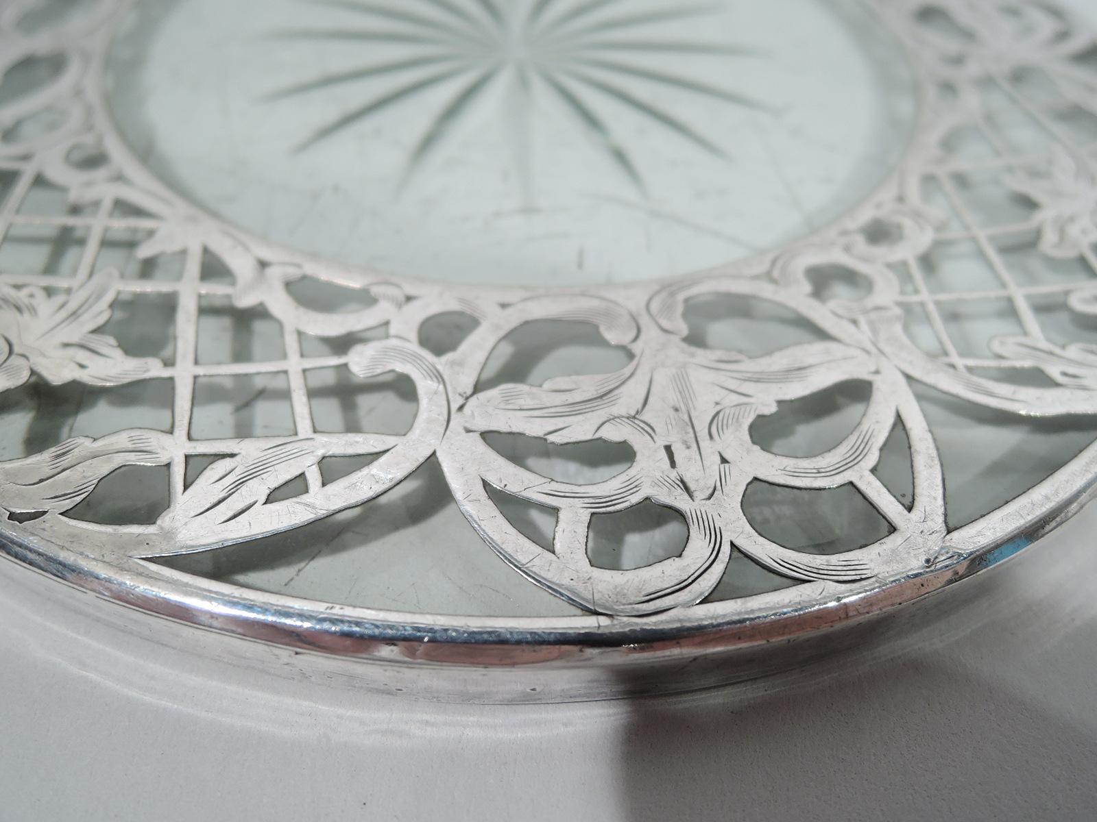 Turn-of-the-century American Art Nouveau glass trivet with engraved silver overlay. Round with star cut to underside bordered by silver overlay trellis and leaves. Glass is clear.