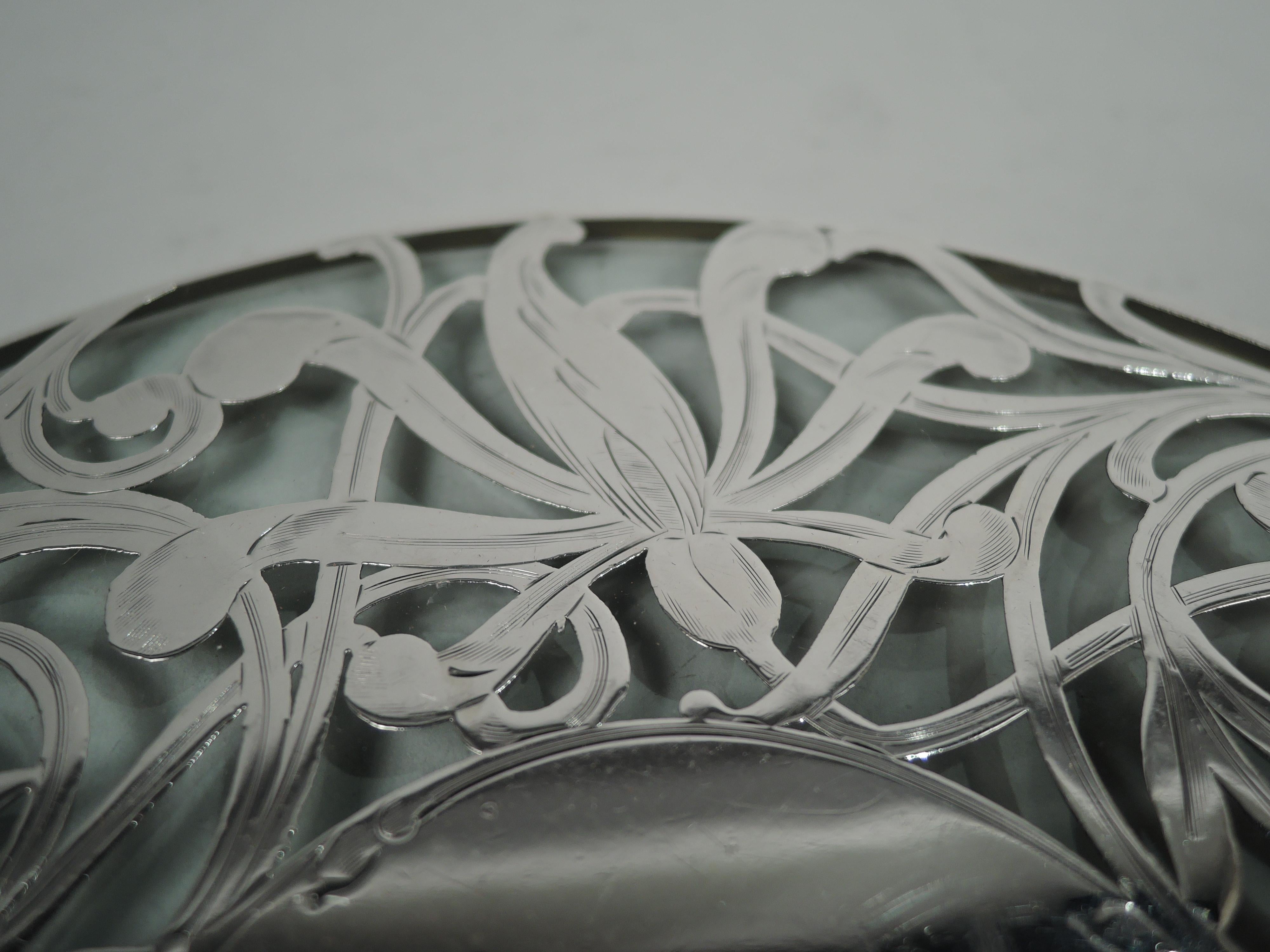 American Art Nouveau glass trivet with engraved silver overlay, ca 1900. Round with straight sides in silver collar. Overlay in form of scrolling and entwined tendrils and distended flowerheads. Center solid and vacant. Glass is clear.