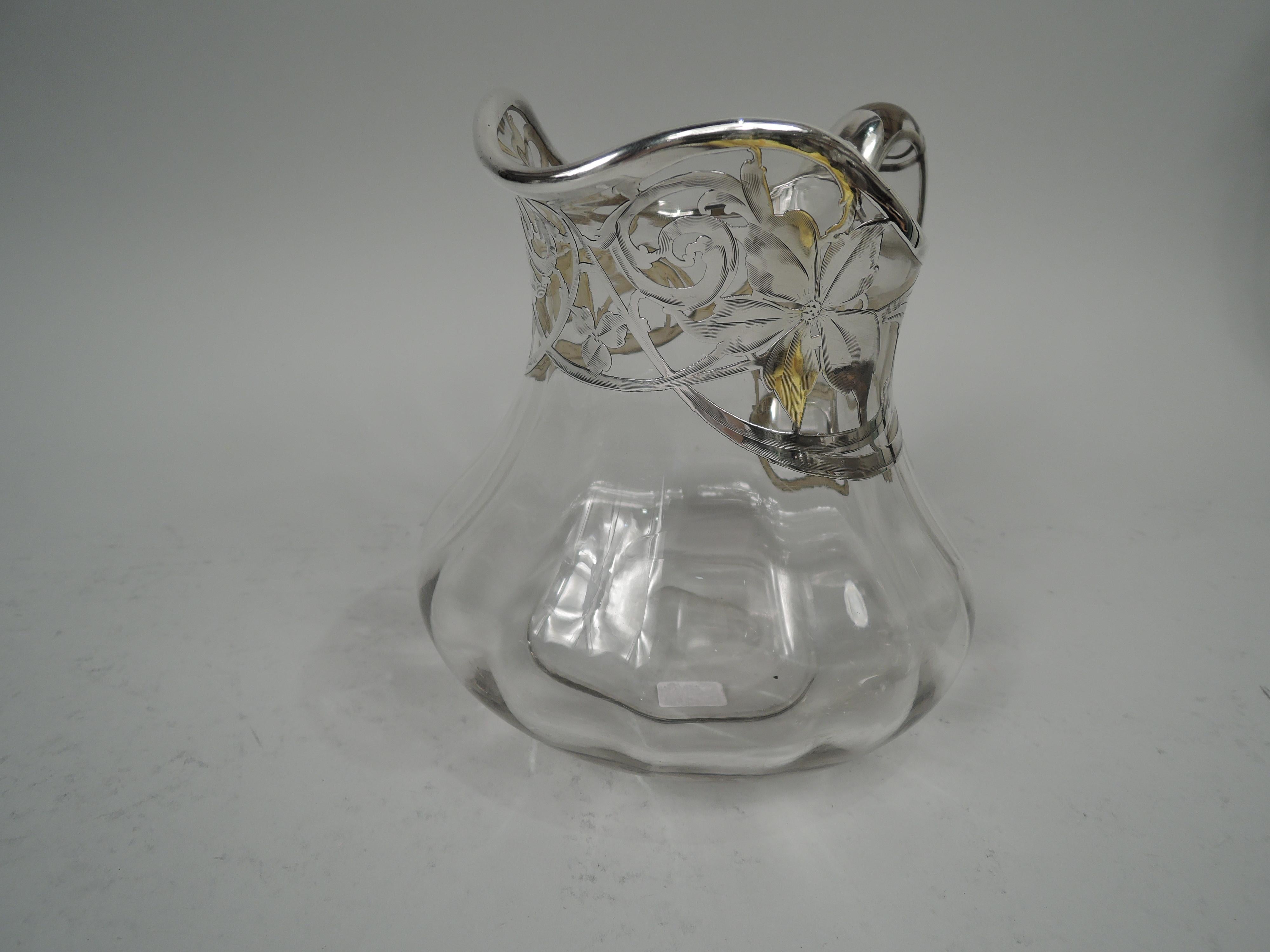 American Art Nouveau glass water pitcher with engraved silver overlay. Low with helmet mouth and spread bottom. Interior lobing and high-looping handles with silver bands. At top overlay in form of leafing scrollwork and flower heads. Glass is