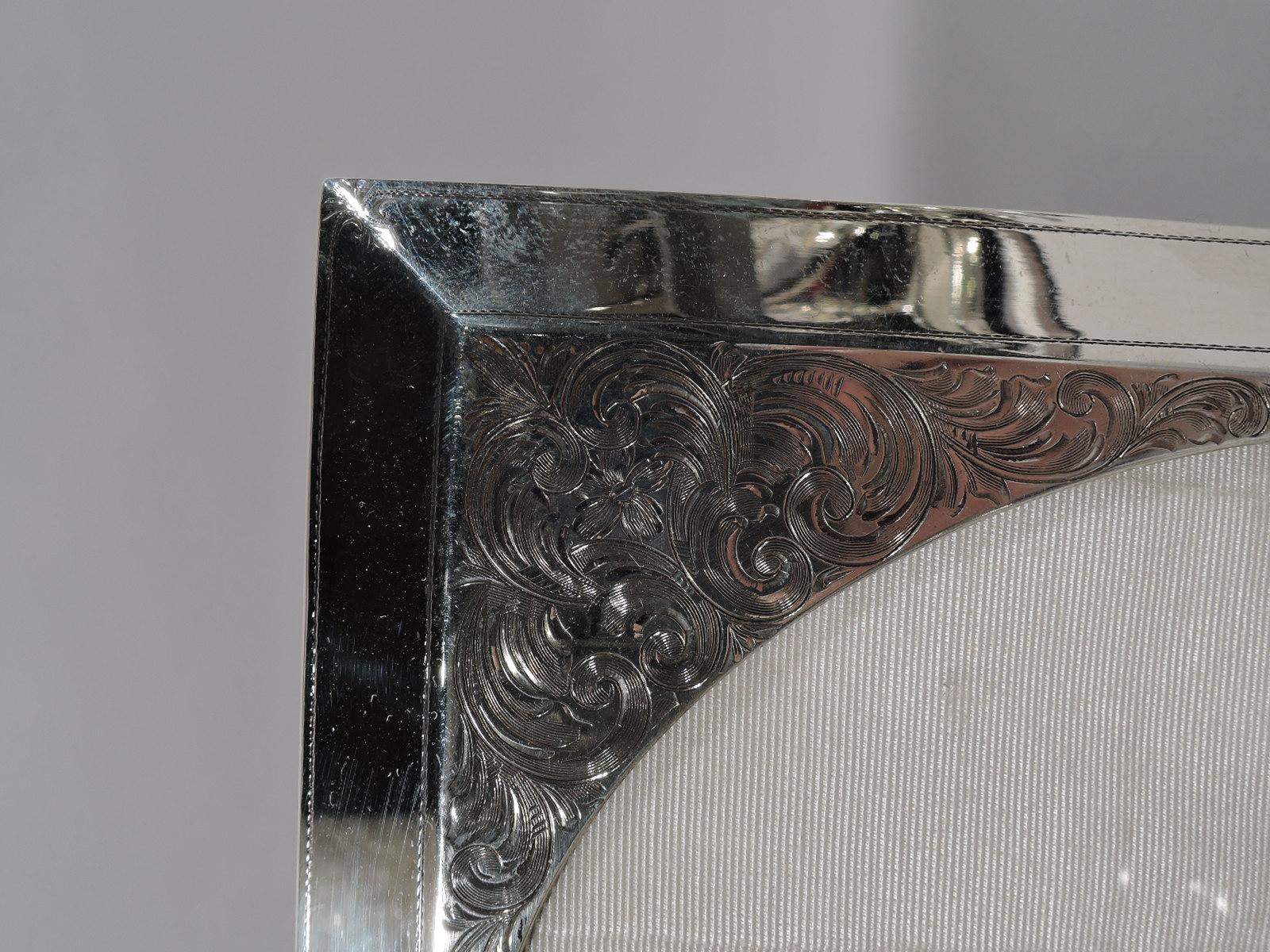 Turn-of-the-century Art Nouveau sterling silver picture frame. Made by William B. Kerr in Newark. Oval window in rectangular surround engraved with dense and fluid scrollwork. Sides canted. With glass, silk lining, and velvet back and hinged