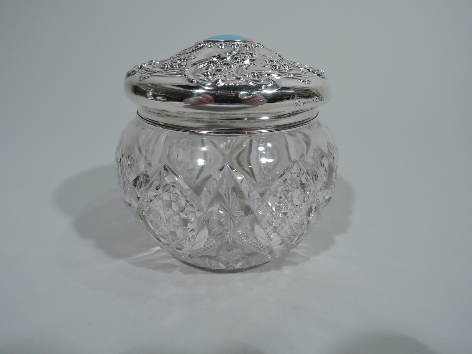 American brilliant-cut glass vanity jar with sterling silver cover. Jar has curved and ornamented sides. Star on underside. Cover has repousse scrolls and flowers and central inset opalescent stone or glass. Hallmark includes no. 1006 and maker’s