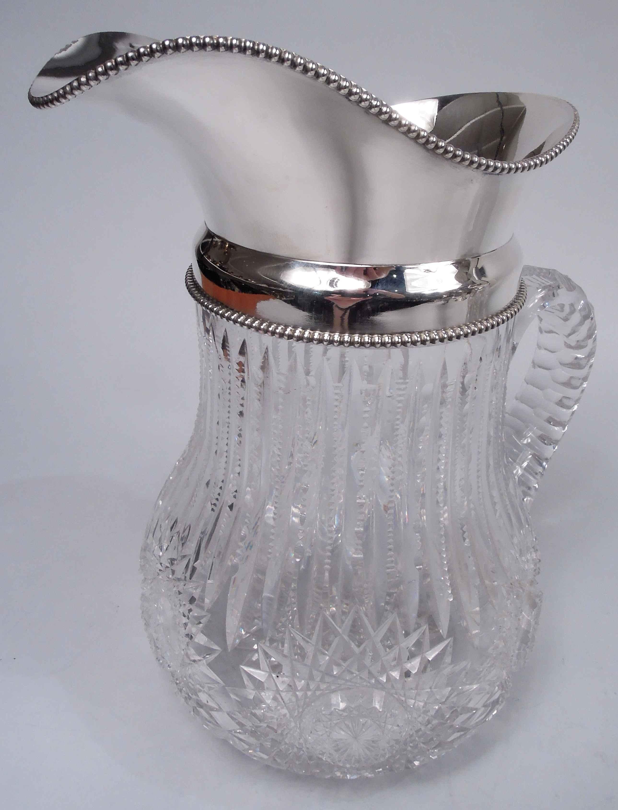 Edwardian Classical brilliant-cut glass water pitcher with sterling silver collar. Made by Wilcox (part of International) in Meriden, Conn., ca 1910. Baluster with c-scroll handle; ornament in form of leaf flutes, ferns, and stars. Beaded collar