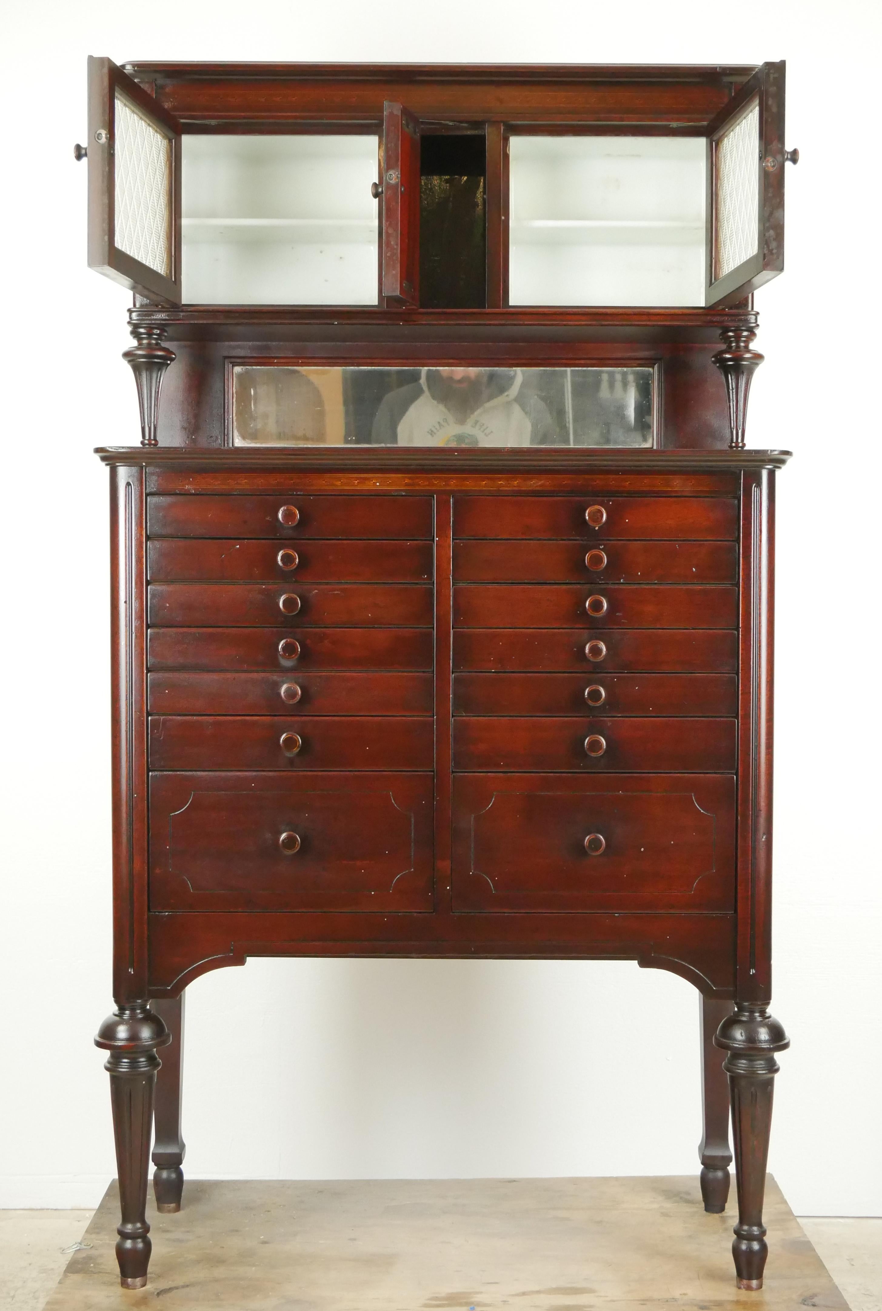 Zahnschrank aus Mahagoni des frühen 20. Jahrhunderts mit 14 Schubladen mit passenden Holzknöpfen, kannelierten Beinen und einer originalen Spiegelrückwand. Der obere Teil hat zwei geprägte Glastüren mit Milchglaseinsätzen. Hergestellt von The