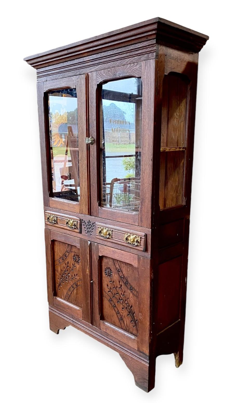 Charmante armoire à tarte en chêne rouge massif, sculptée à la main, avec un décor folklorique de fleurs et de feuilles, comportant deux tiroirs avec des poignées en laiton d'origine, une étagère en bois derrière les portes en verre supérieures et