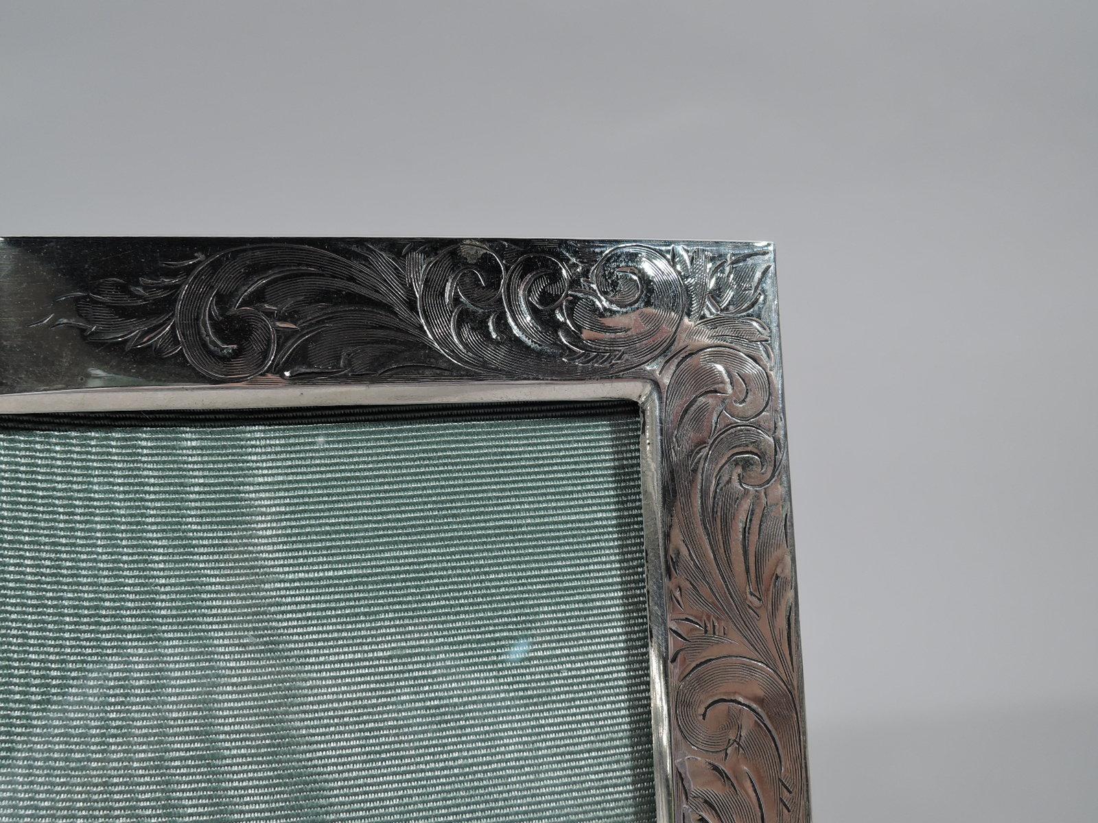 Edwardian Art Nouveau sterling silver picture frame, ca 1910. Rectangular window in flat surround with densely engraved scrolls and flowers; sides plain. Top rail center vacant. With glass, silk lining, and velvet back and hinged easel support for