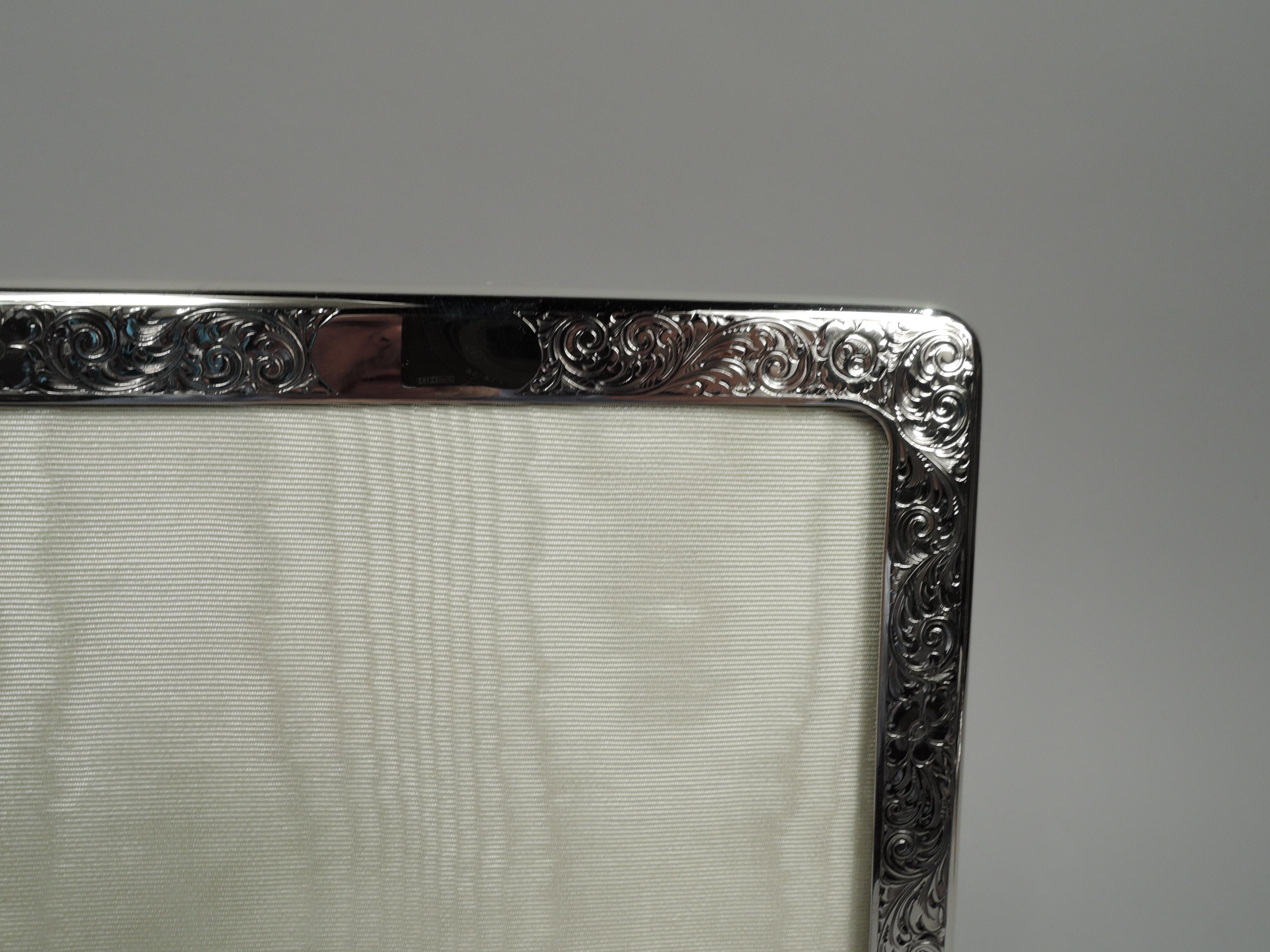 Edwardian Art Nouveau sterling silver picture frame, ca 1910. Rectangular window in surround with curved corners. Front engraved with dense leafing scrollwork and flowers; tubular cartouche (vacant). Sides plain. With glass, silk lining, and velvet