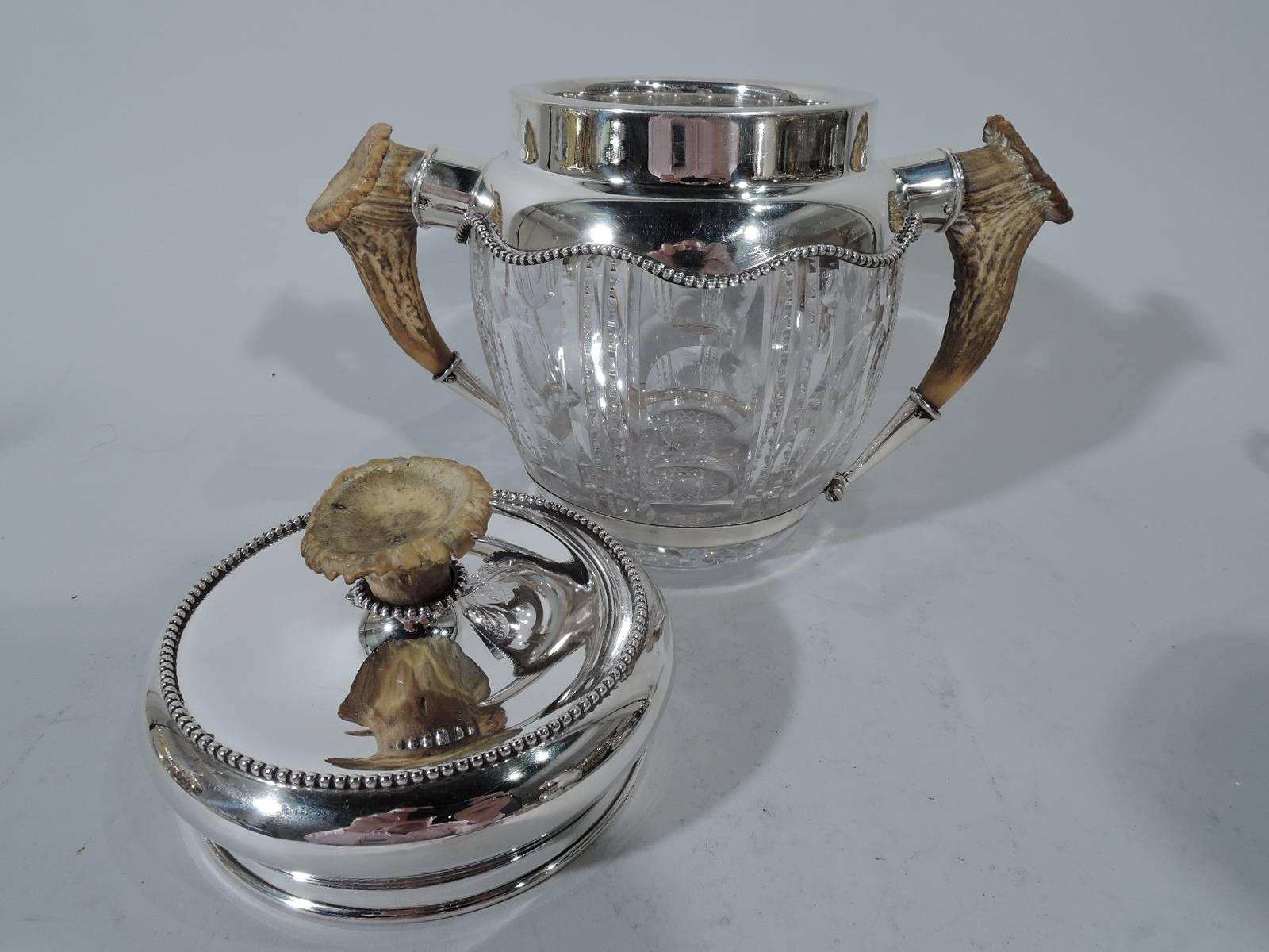 Edwardian cut-glass tobacco jar with sterling silver mounts and cover. Made by Redlich & Co. in New York. Jar has curved sides with vertical ornament: circles alternate with notched double flutes, and antler side handles in mounts. Short neck has