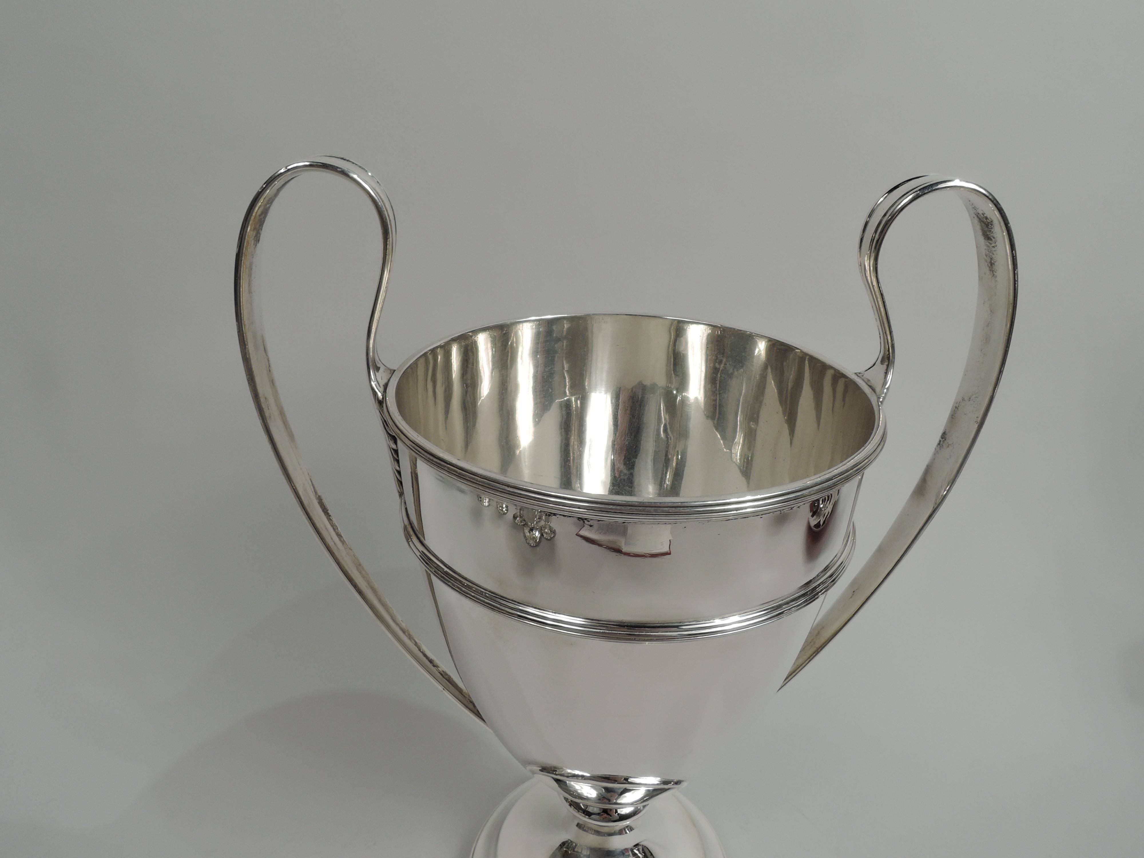 Edwardian Classical sterling silver trophy cup. Made by Howard & Co. in New York, ca 1910. Traditional amphora urn: Girdled ovoid bowl on stepped and domed foot. High-looping side handles with triangular leaf mounts. Reeded rims. Traditional