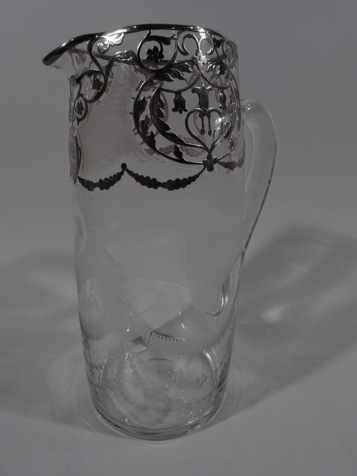 American Edwardian Regency claret jug, ca 1915. Curved cylinder with high-looping handle in clear glass with acid-etched ornament: stylized garland and flowers, and shaped fish-scale surrounding silver overlay in form of leafy scrolls and garland.