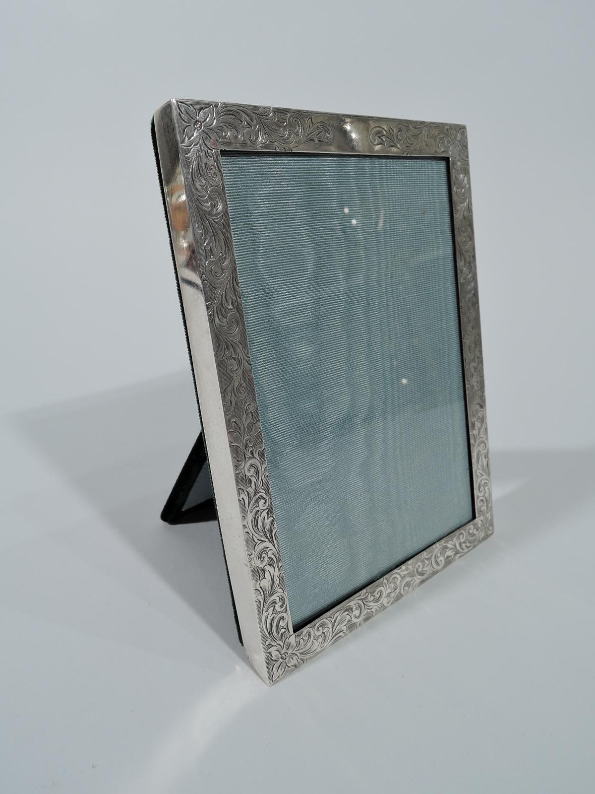 Edwardian sterling silver picture frame. Rectangular window and flat border engraved with fluid scrollwork and flowers. Top vacant. With glass, silk lining, and velvet back and hinged support. For portrait (vertical) display. Hallmarked IN Deitsch,