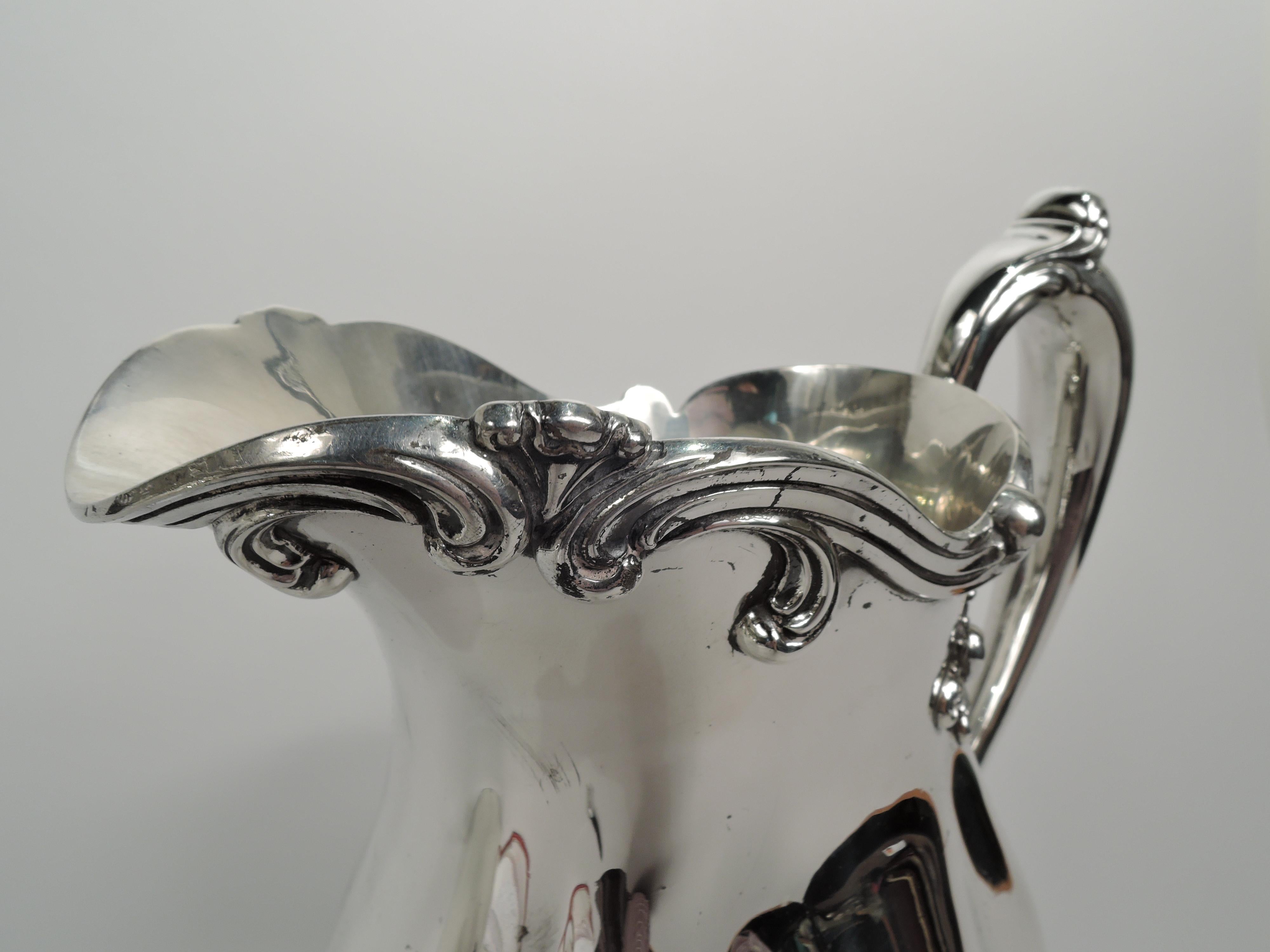 Edwardian sterling silver water pitcher. Made by Frank M. Whiting Co. in North Attleboro, Mass., ca 1910. Lobed and oval body on short and scalloped foot. Capped and scroll-mounted high-looping handle. Scrolled helmet mouth with applied rim. Pretty