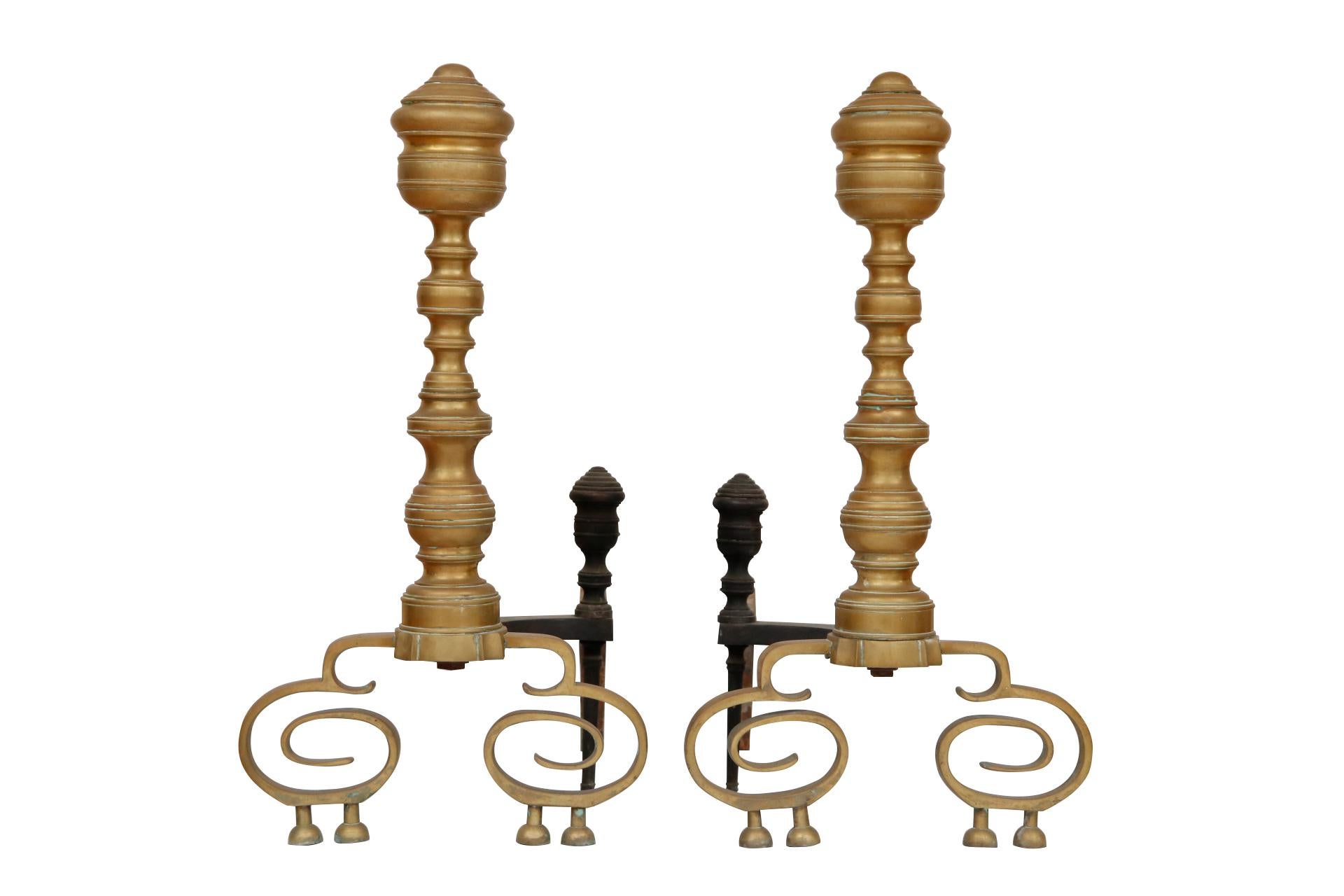 A pair of American Empire brass andirons circa 1830. Columns are cast to give the look of turned balusters with large urn finials. Matching log stops decorate wrought iron dog legs. The andirons stand on scrolled legs each finished with ball feet.