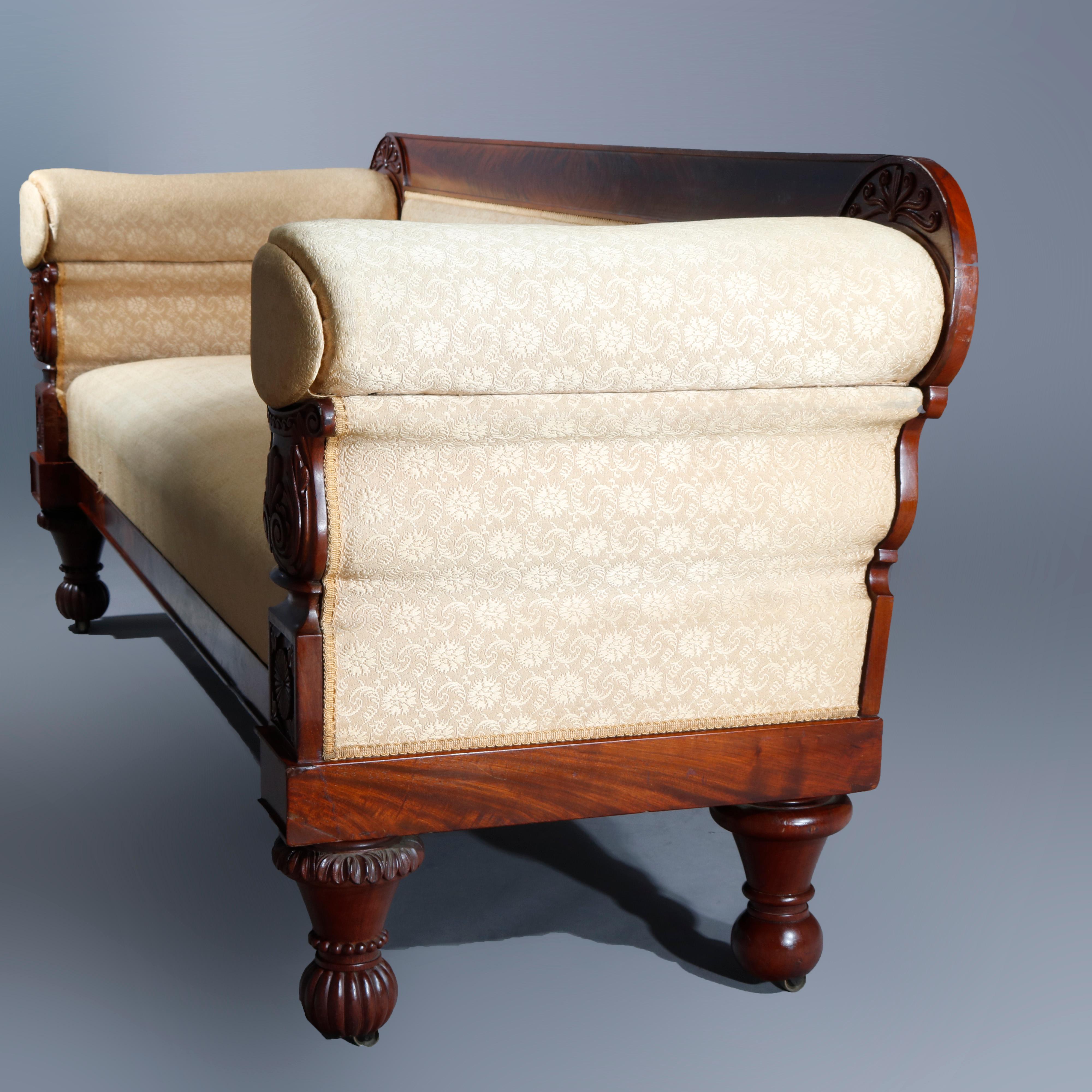 An antique American Empire sofa in the manner of Anthony Quervelle offers flame mahogany frame with straight crest flanked by caved stylized scrolled foliate reserves over upholstered back, arms and seat with arms having carved urn form supports and