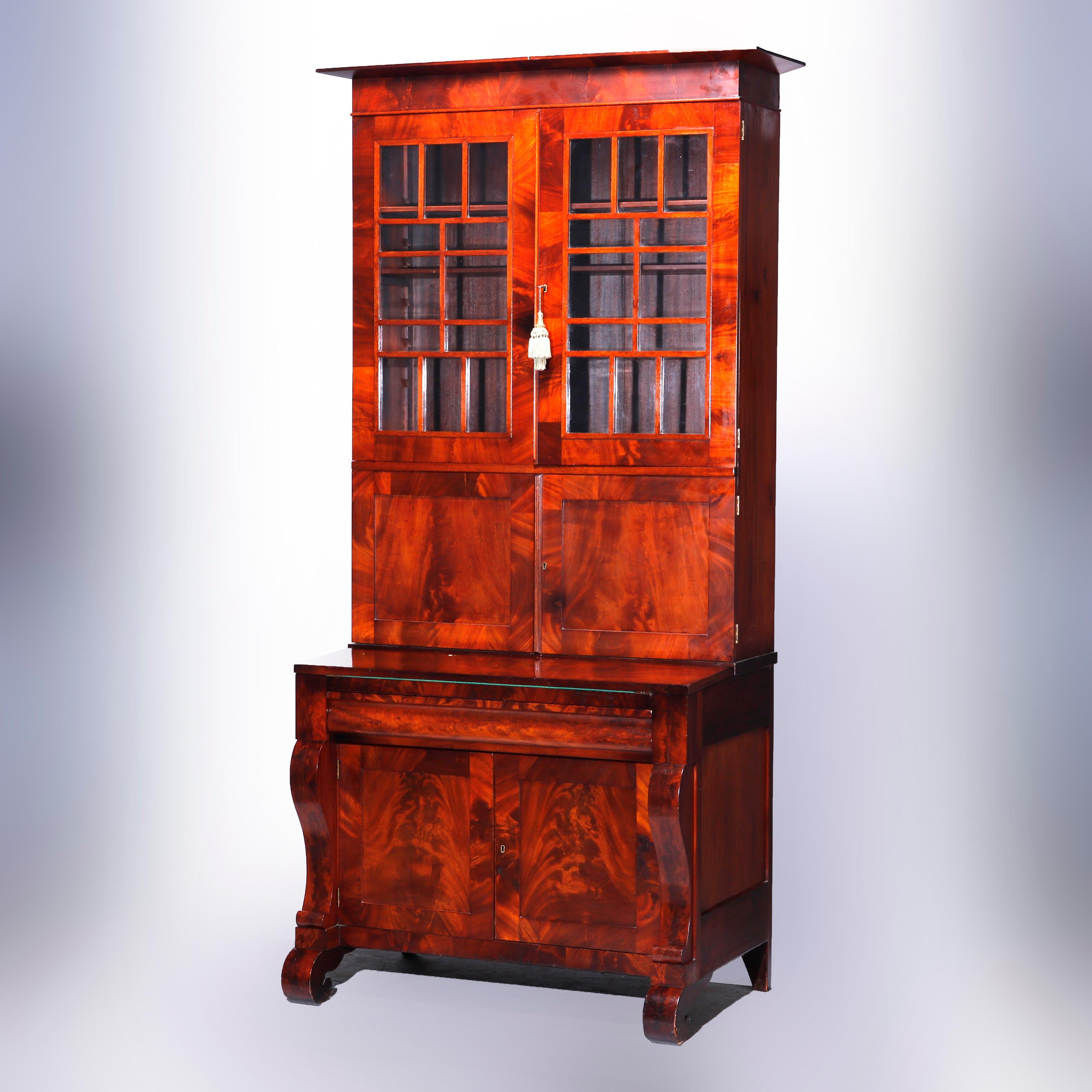 An antique American Empire secretary offers flame mahogany construction with upper bookcase having mullioned glass doors (patterning reminiscent of Fran Lloyd Wright) opening to shelved interior and over double blind door lower cabinet opening to