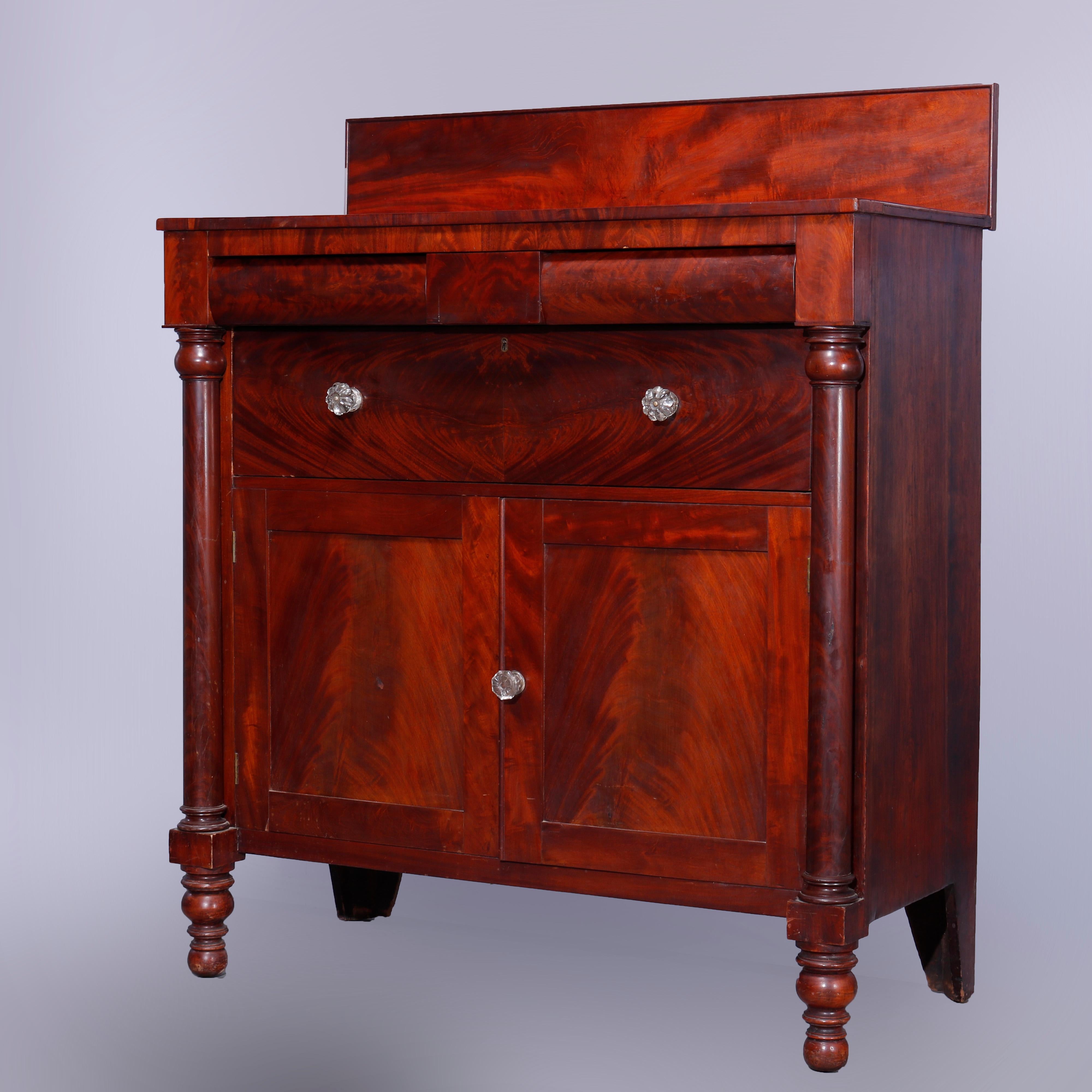 An antique American Empire classical grecian sideboard offers flame mahogany construction with backsplash surmounting case with convex double drawers over deep frieze drawer and double door lower cabinet with flanking Doric column supports, raised