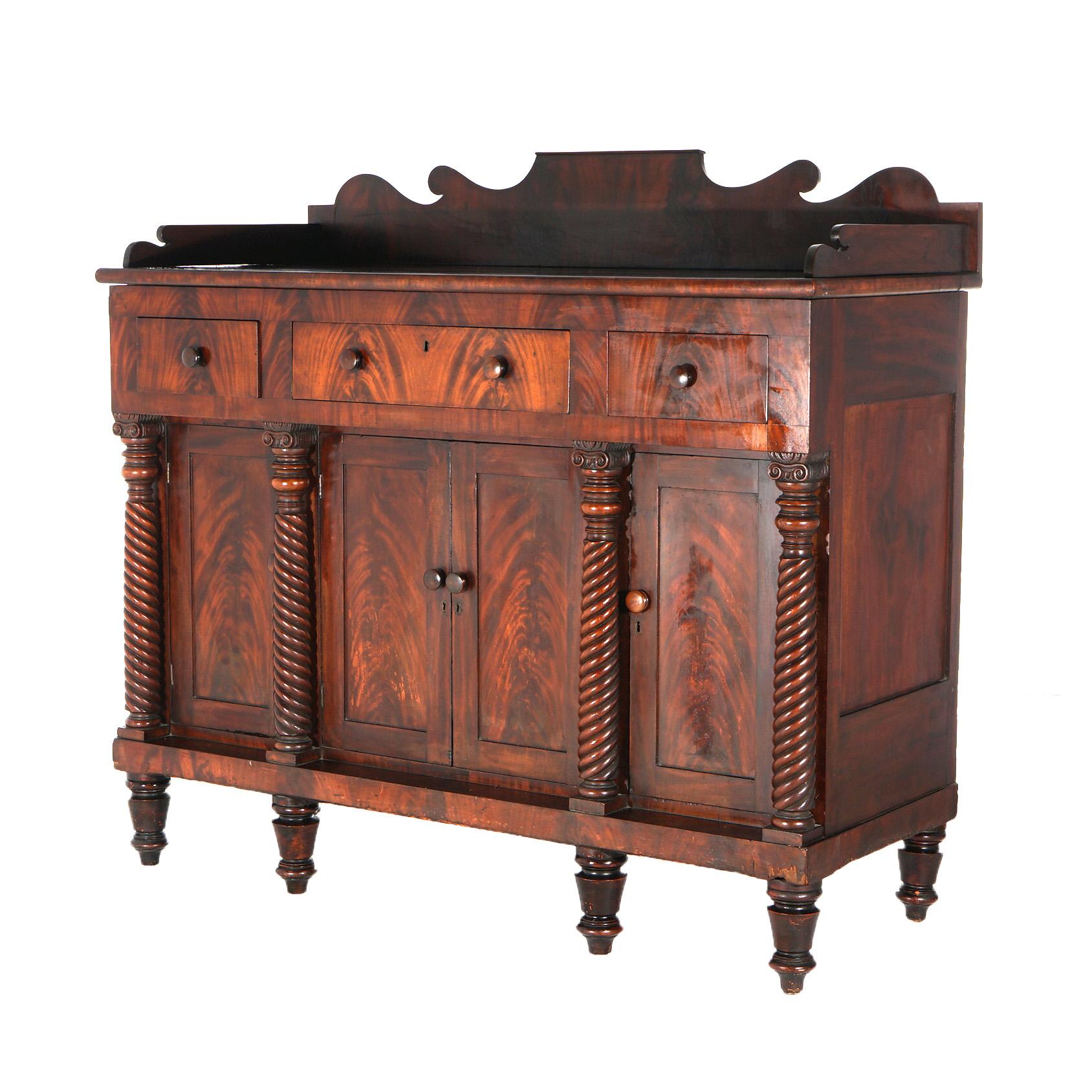 An antique American Empire Classical Greco sideboard offers flame mahogany paneled construction with scroll for backsplash over case with upper drawers over lower cabinets flanked by rope twist Corinthian column supports, c1860

Measures - 53.5