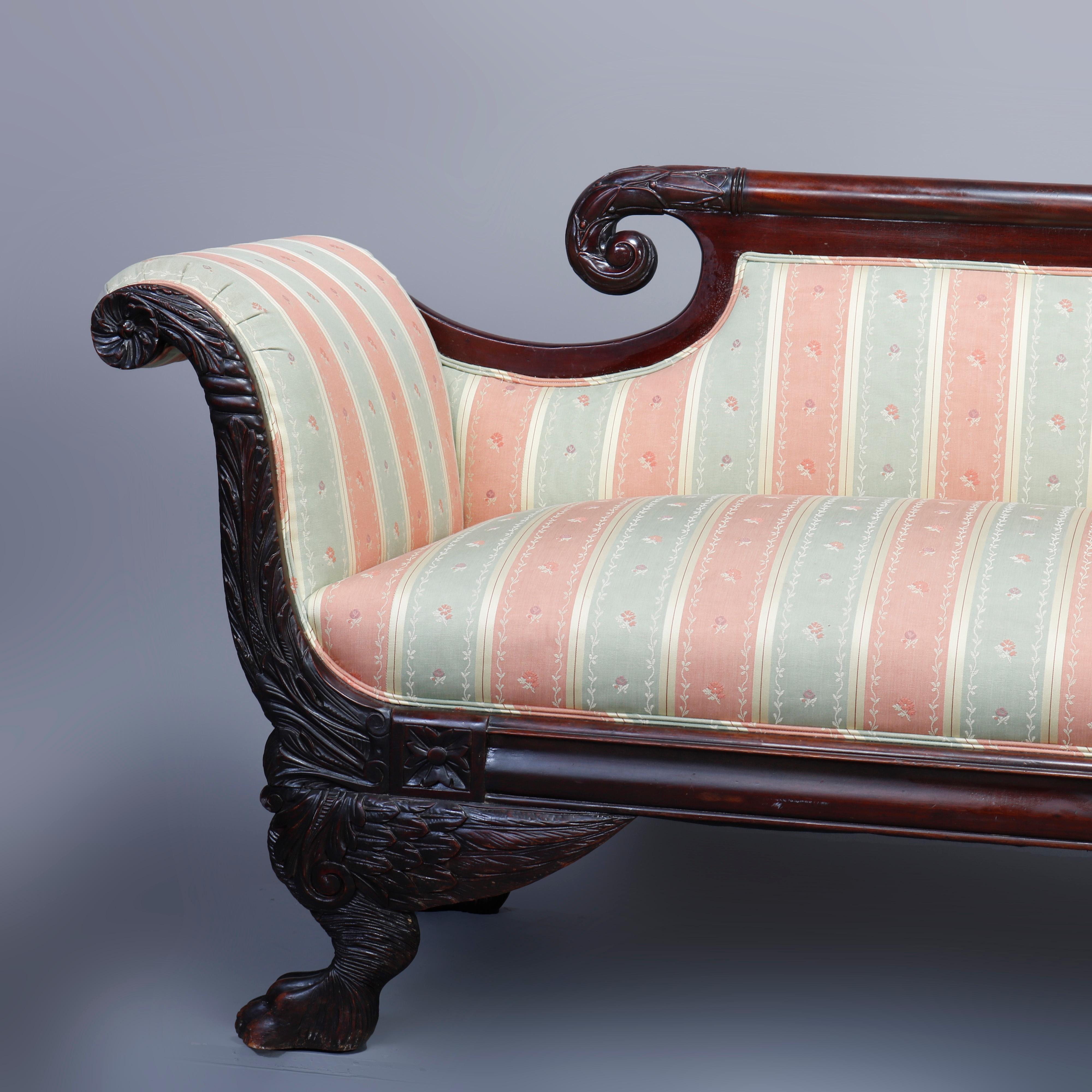 An antique American Empire classical sofa offers mahogany frame with carved scrolled crest surmounting upholstered back, seat and scroll arms having foliate carved decoration, raised on carved paw feet with scroll and feathered wings, 19th