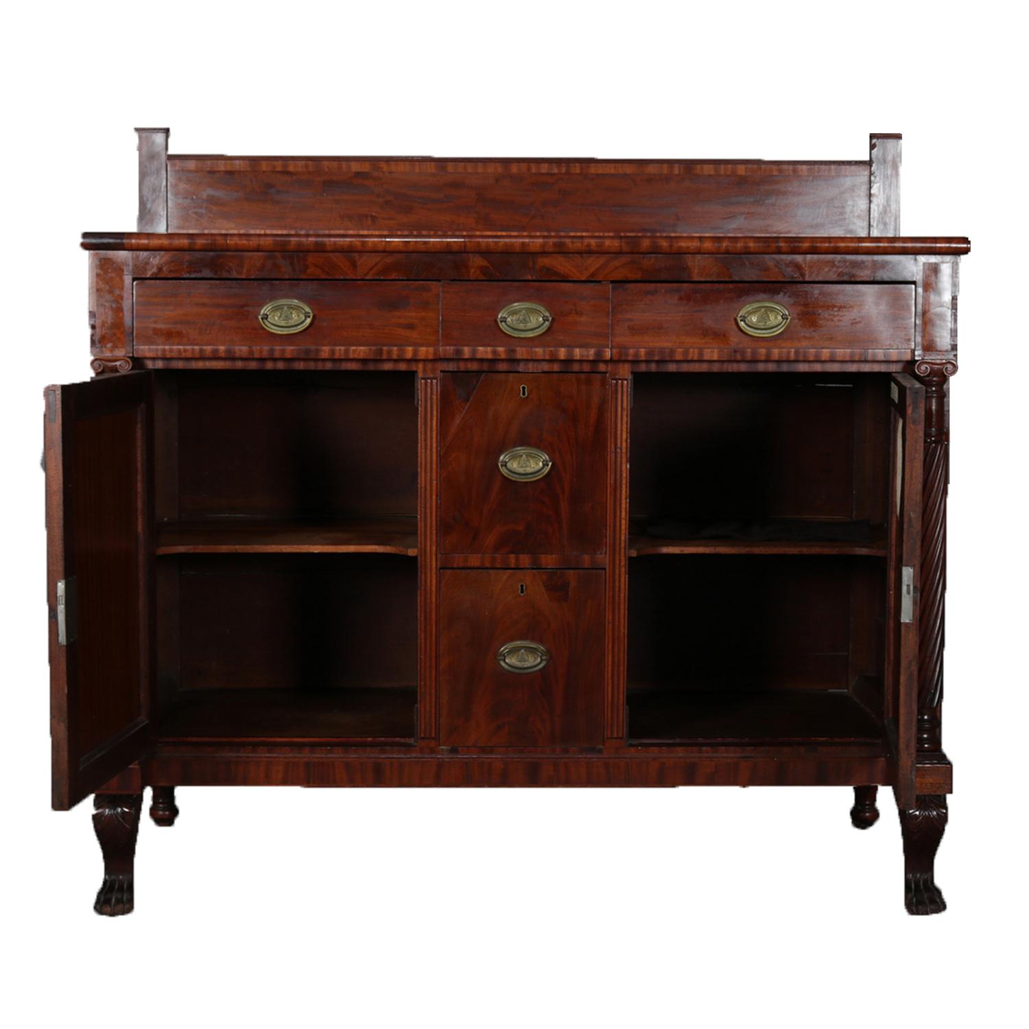 An antique American Empire mahogany sideboard features flame mahogany construction with crossbanded backsplash surmounting case having lined silver drawer, central drawer and velvet lined divided drawer over two stacked central drawers flanked by