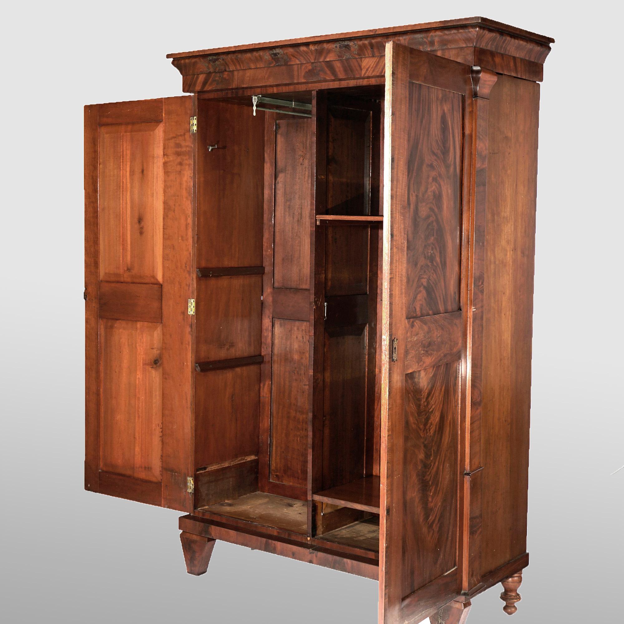 An antique American Empire armoire offers paneled flame mahogany construction with case having double doors opening to a divided interior with shelves and a clothes bar, flanking stylized column supports and raised on square and tapered squat legs,